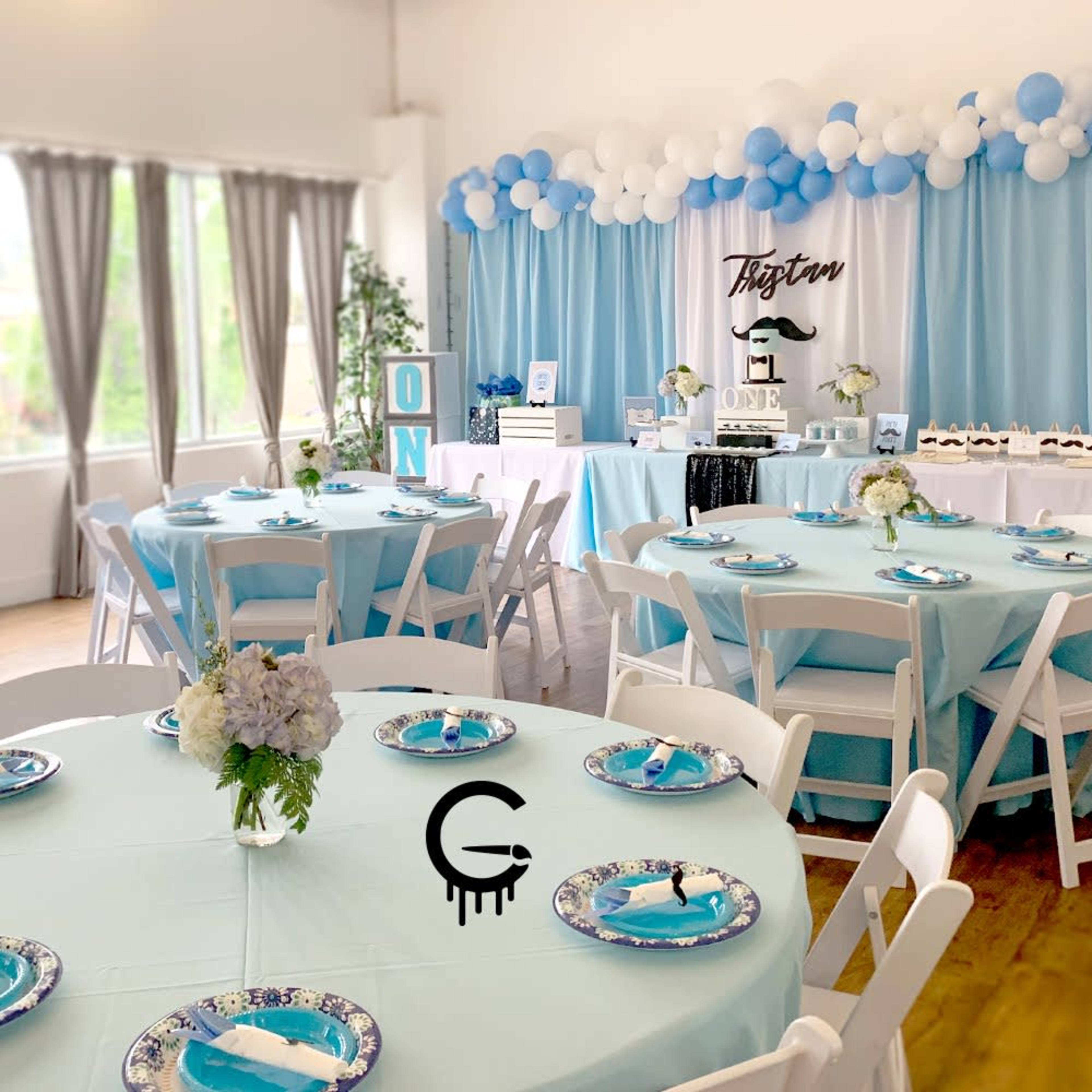 A decorated event space with tables set in light blue and white, featuring floral centerpieces and a festive backdrop with balloons and a cake display.