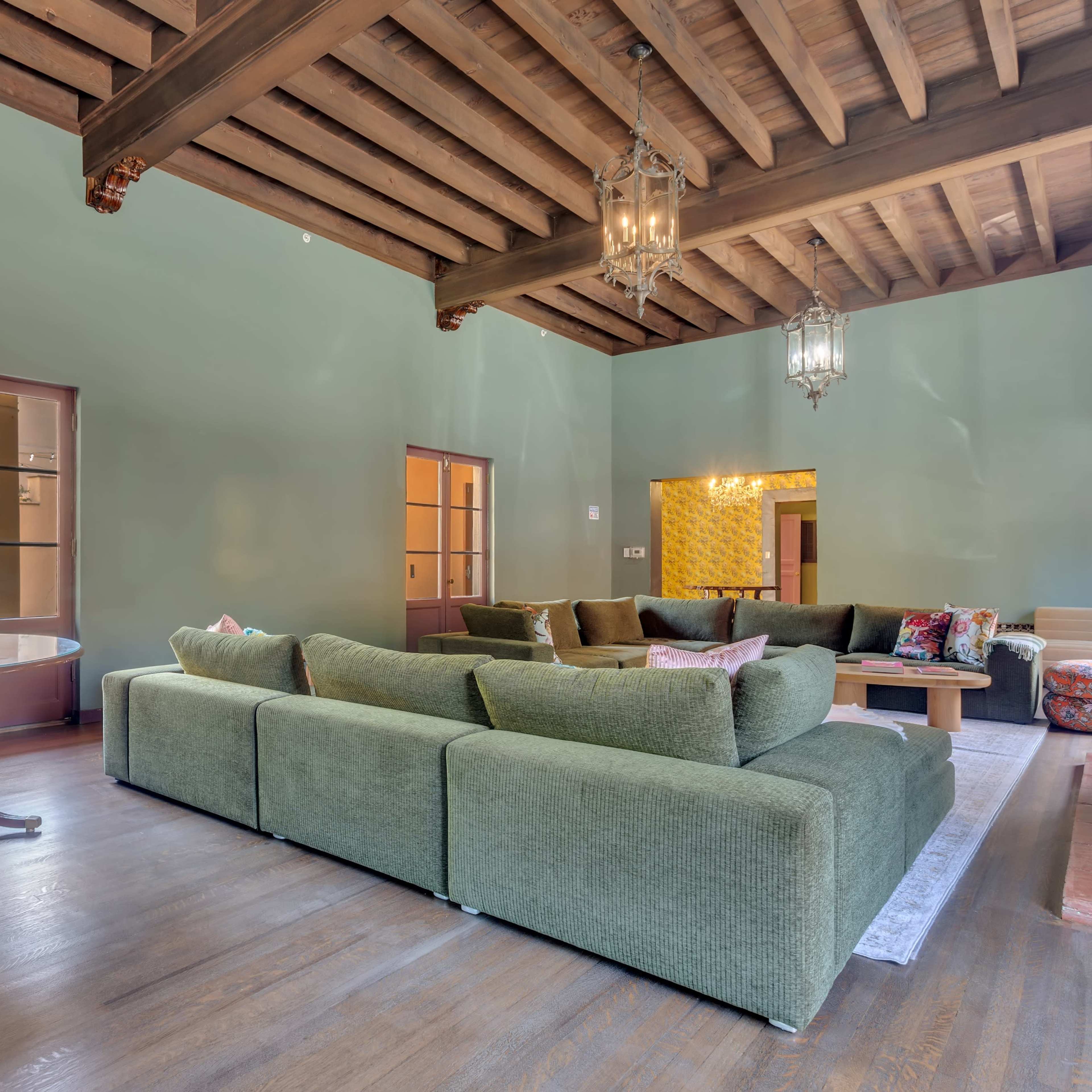 A spacious living room features a large green sectional sofa, wooden ceiling beams, and multiple windows with natural light.