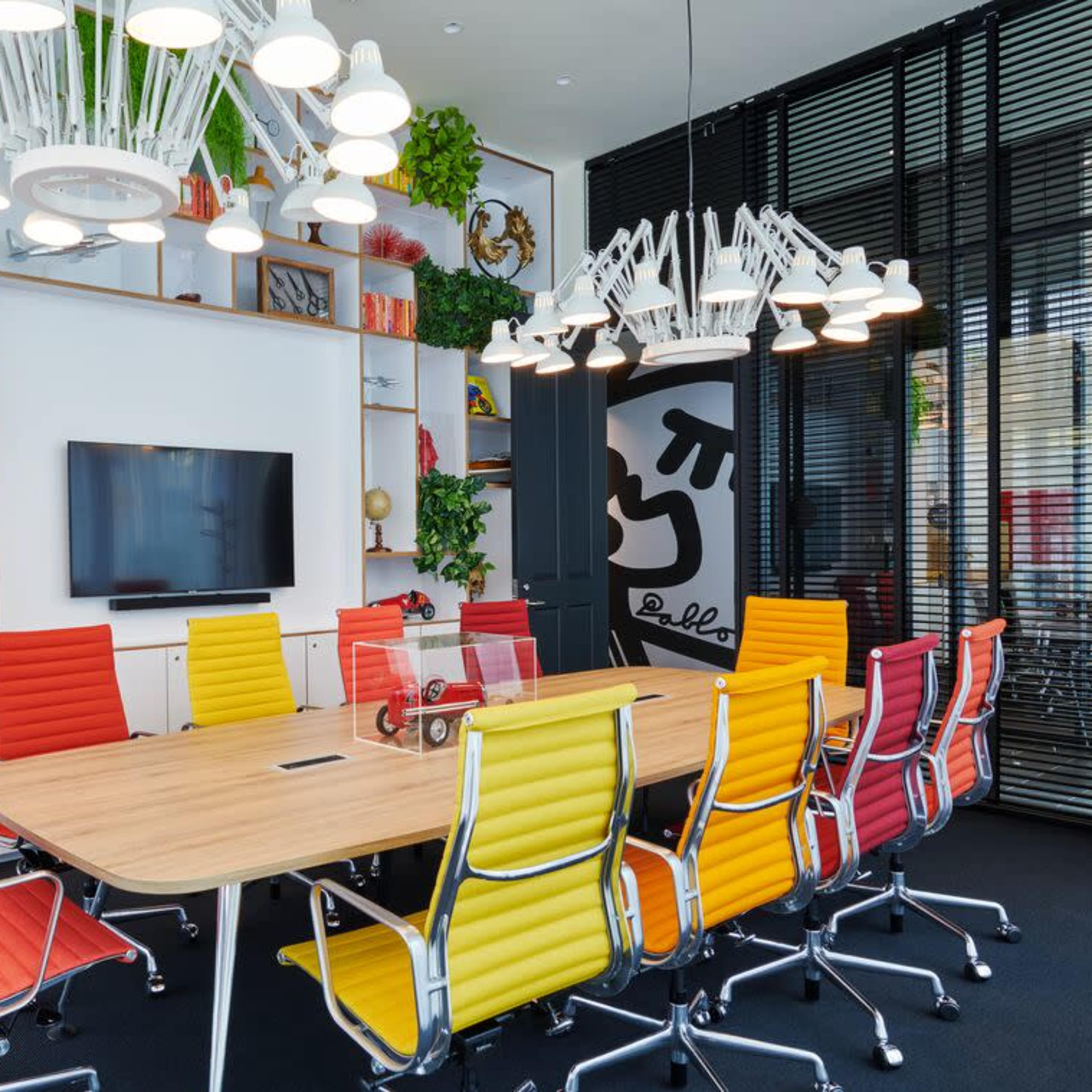 A bright conference room features a large wooden table surrounded by colorful chairs, with a wall-mounted TV and decorative plants.