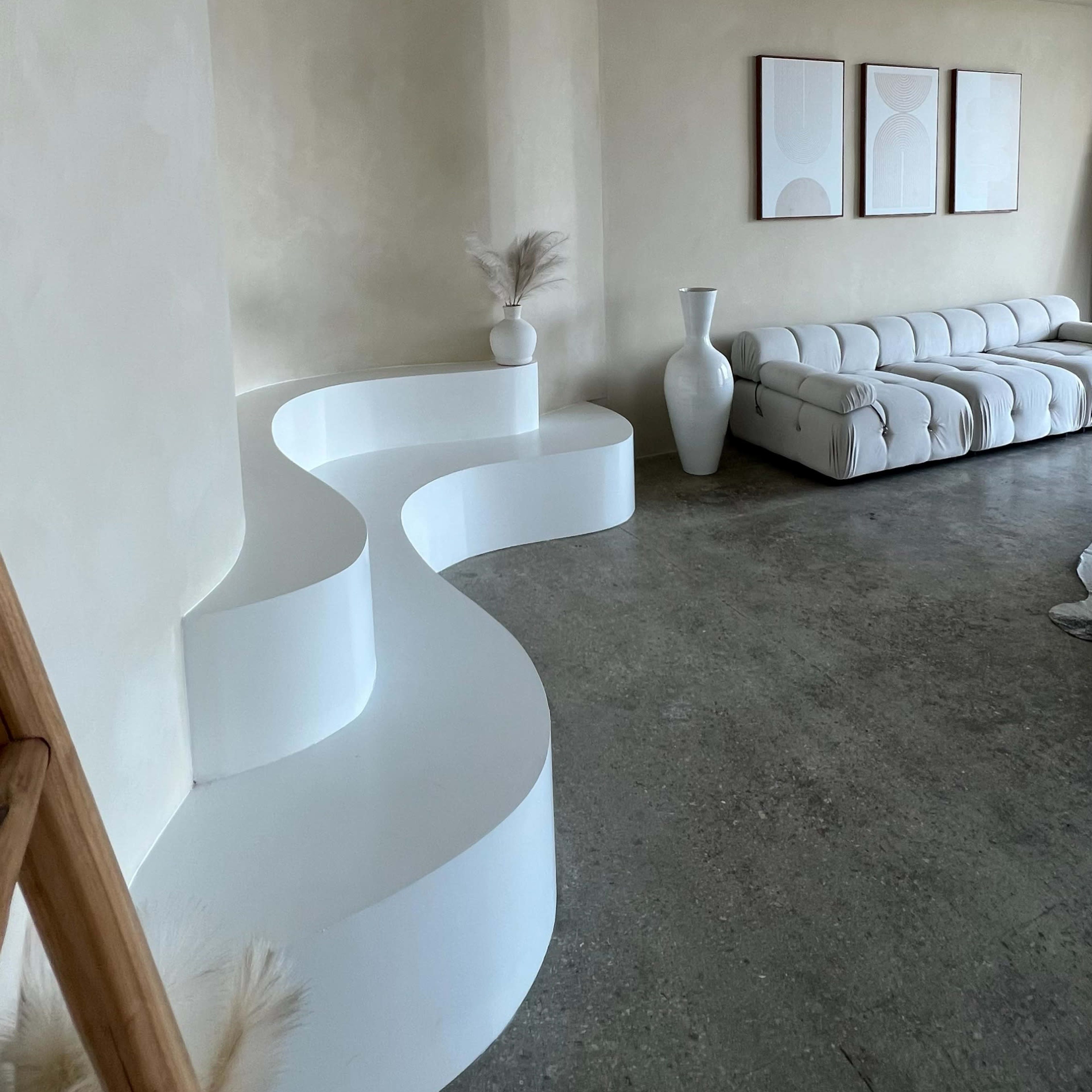 A minimalistic living room featuring a curved white seating area, a white vase, and three framed artworks on the wall.