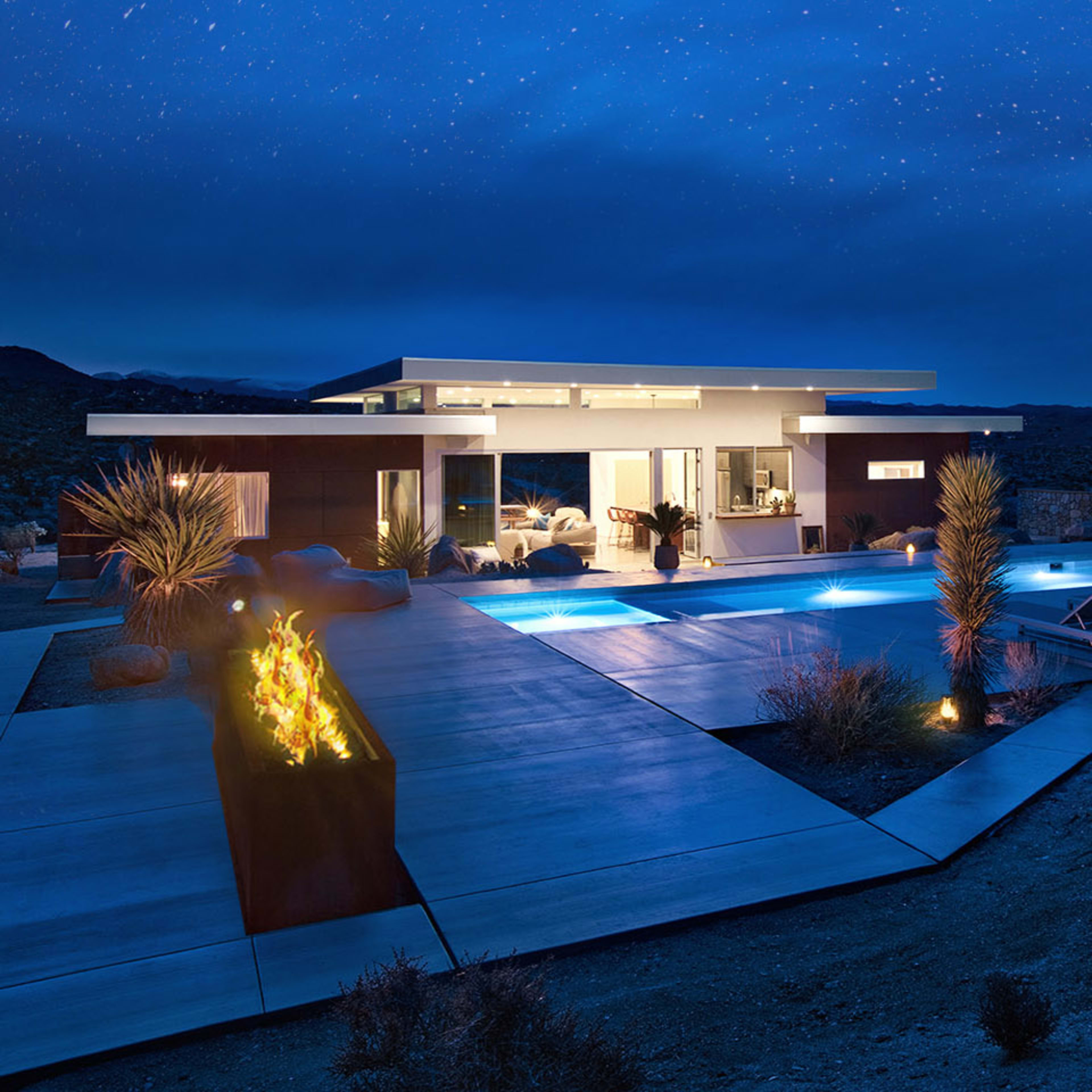 A modern home with a pool and outdoor seating is illuminated under a starry night sky in a desert landscape.