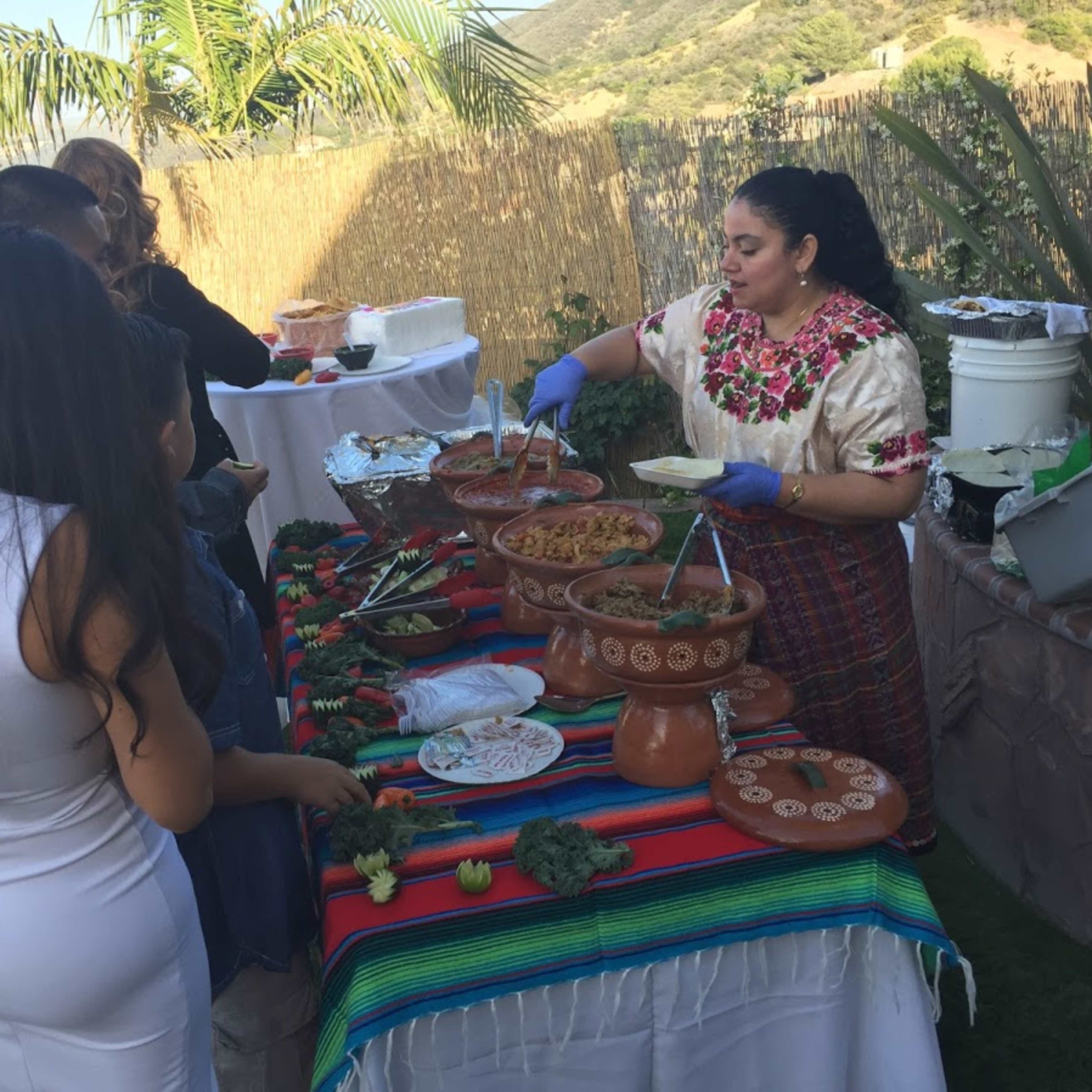 A woman in traditional clothing serves food from clay pots at an outdoor gathering.