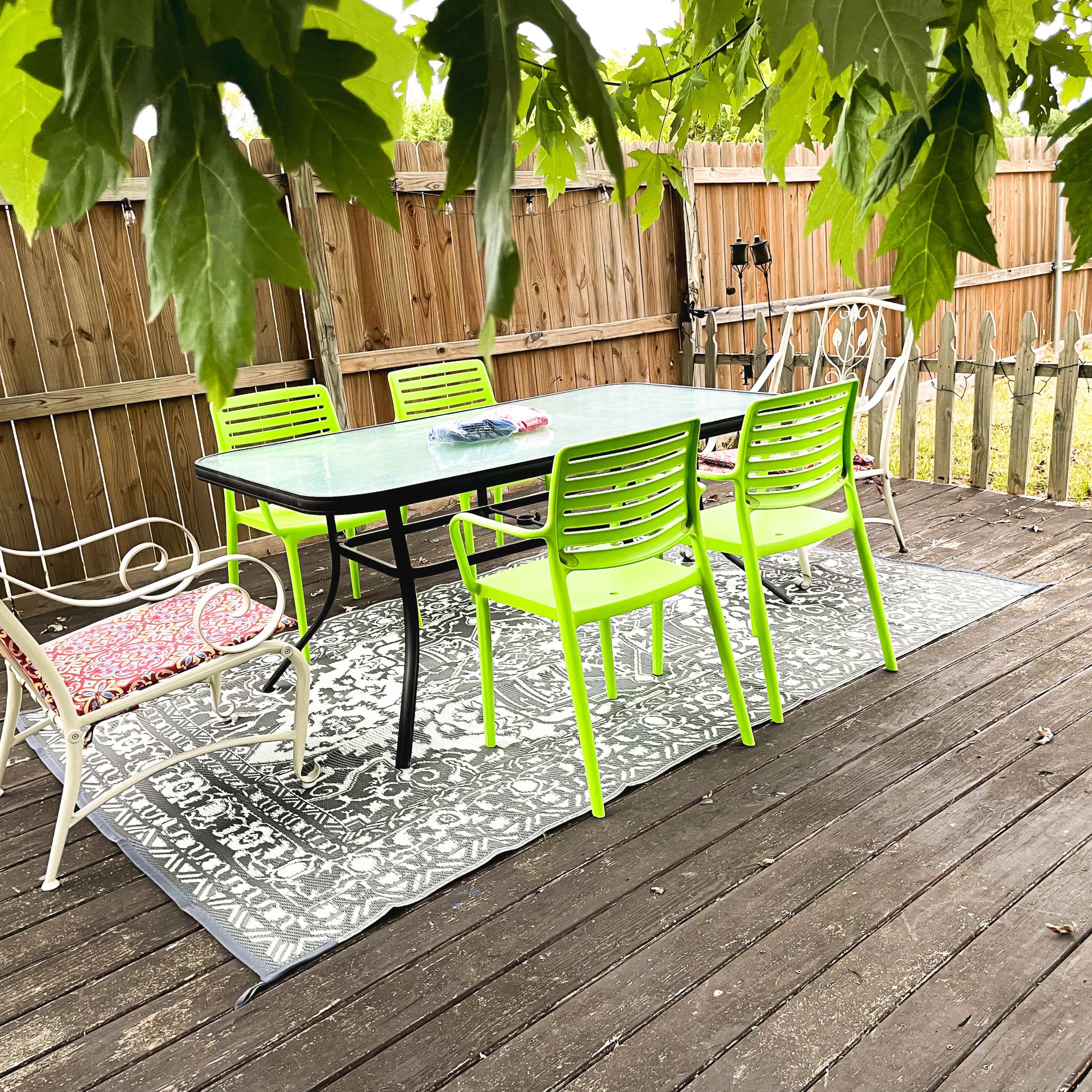 A patio area with a table and six chairs, surrounded by wooden fencing and covered by overhanging leaves.