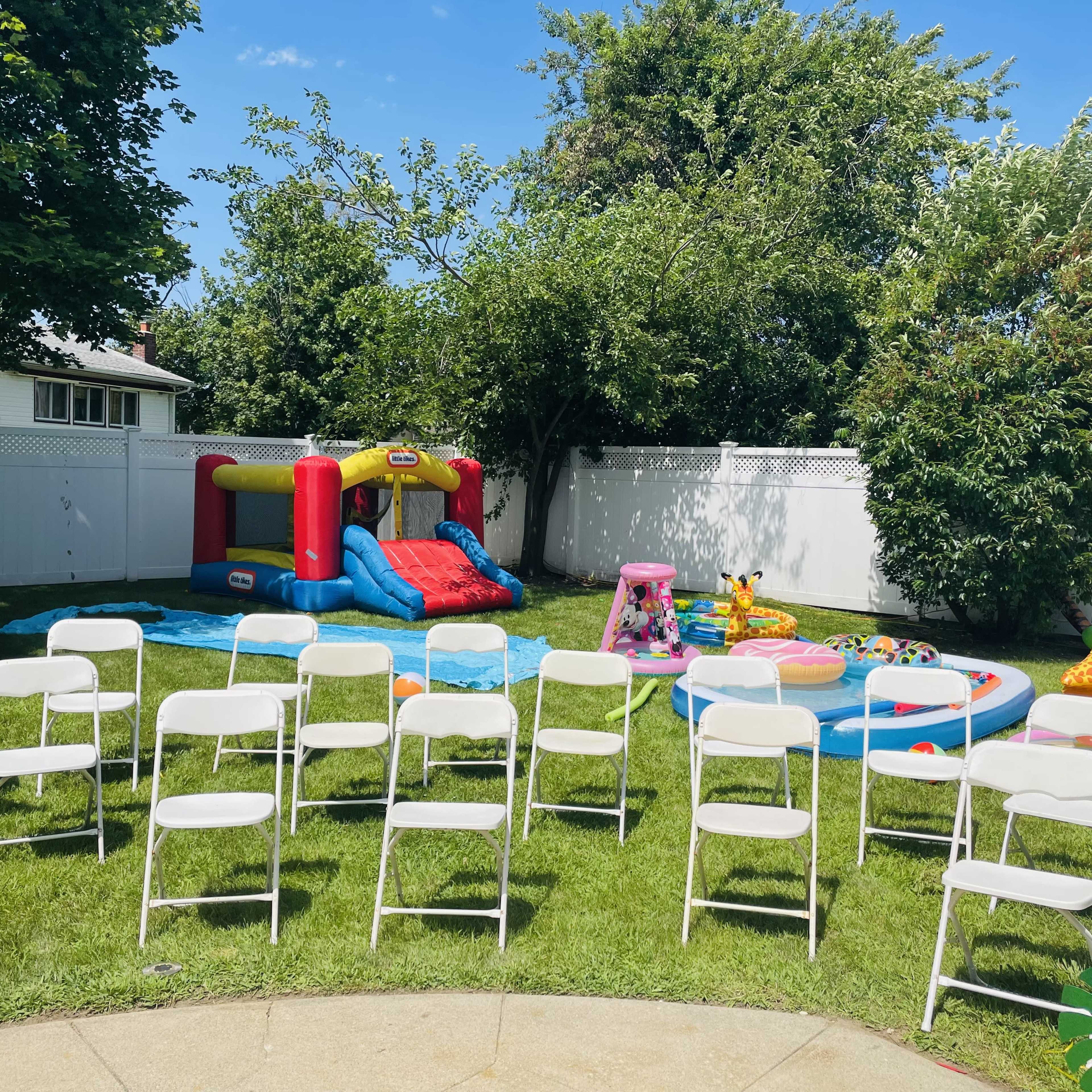 A backyard features an inflatable bounce house, a water slide, and various inflatable toys surrounded by white folding chairs on a grassy area.