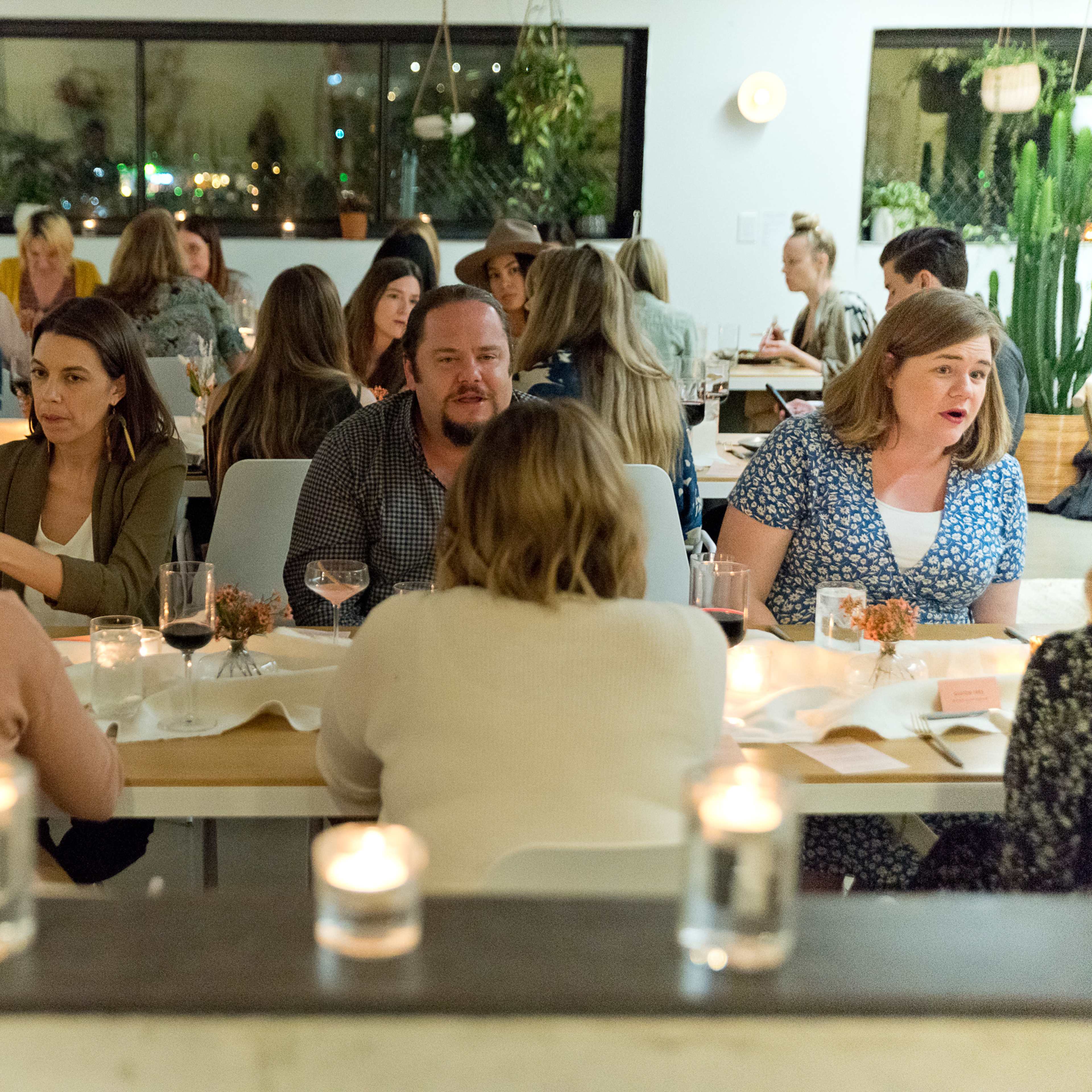 A group of people sits at tables in a well-lit restaurant, engaged in conversation while candles flicker on the tables.