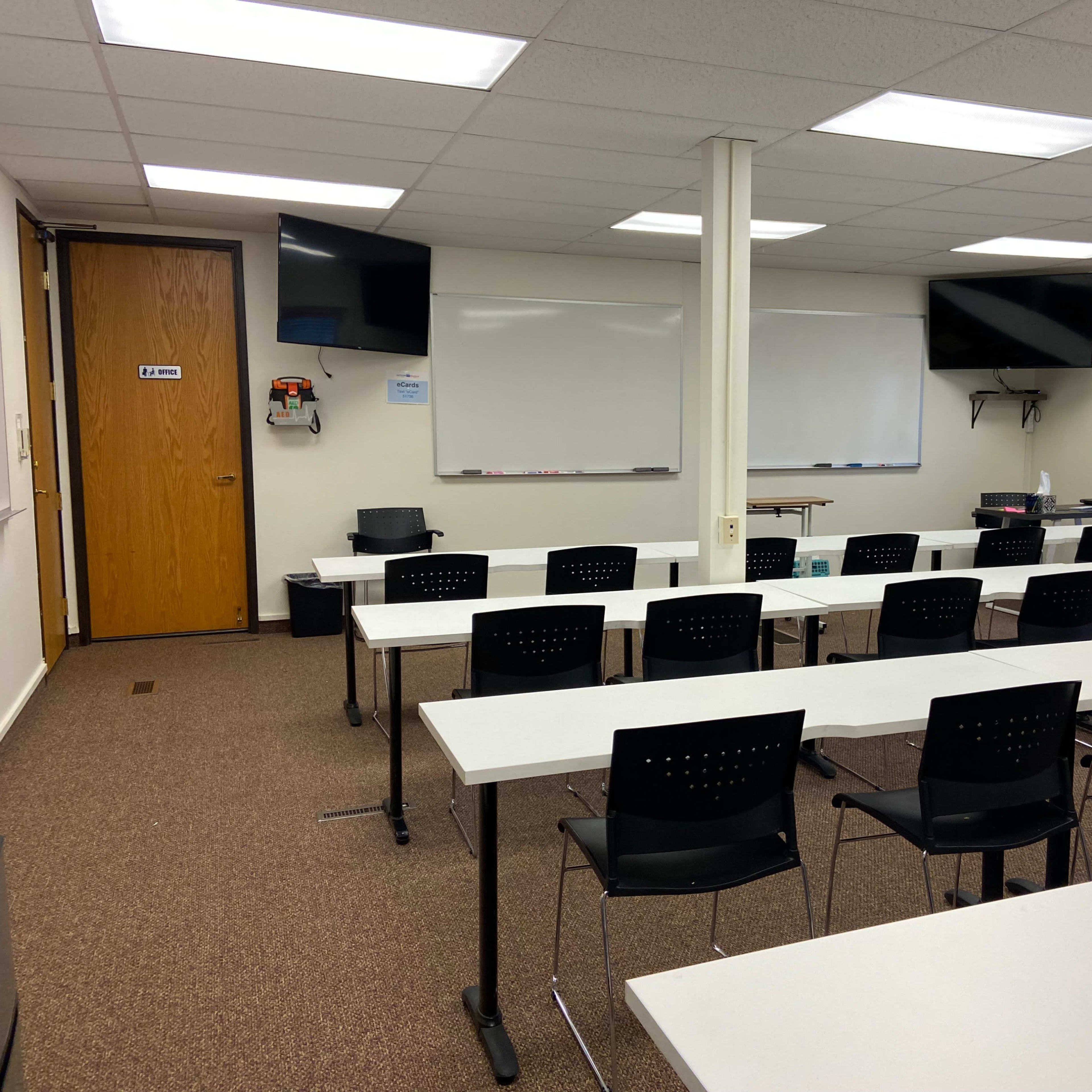 A classroom features several rows of white tables with black chairs, two large whiteboards, and two wall-mounted screens.