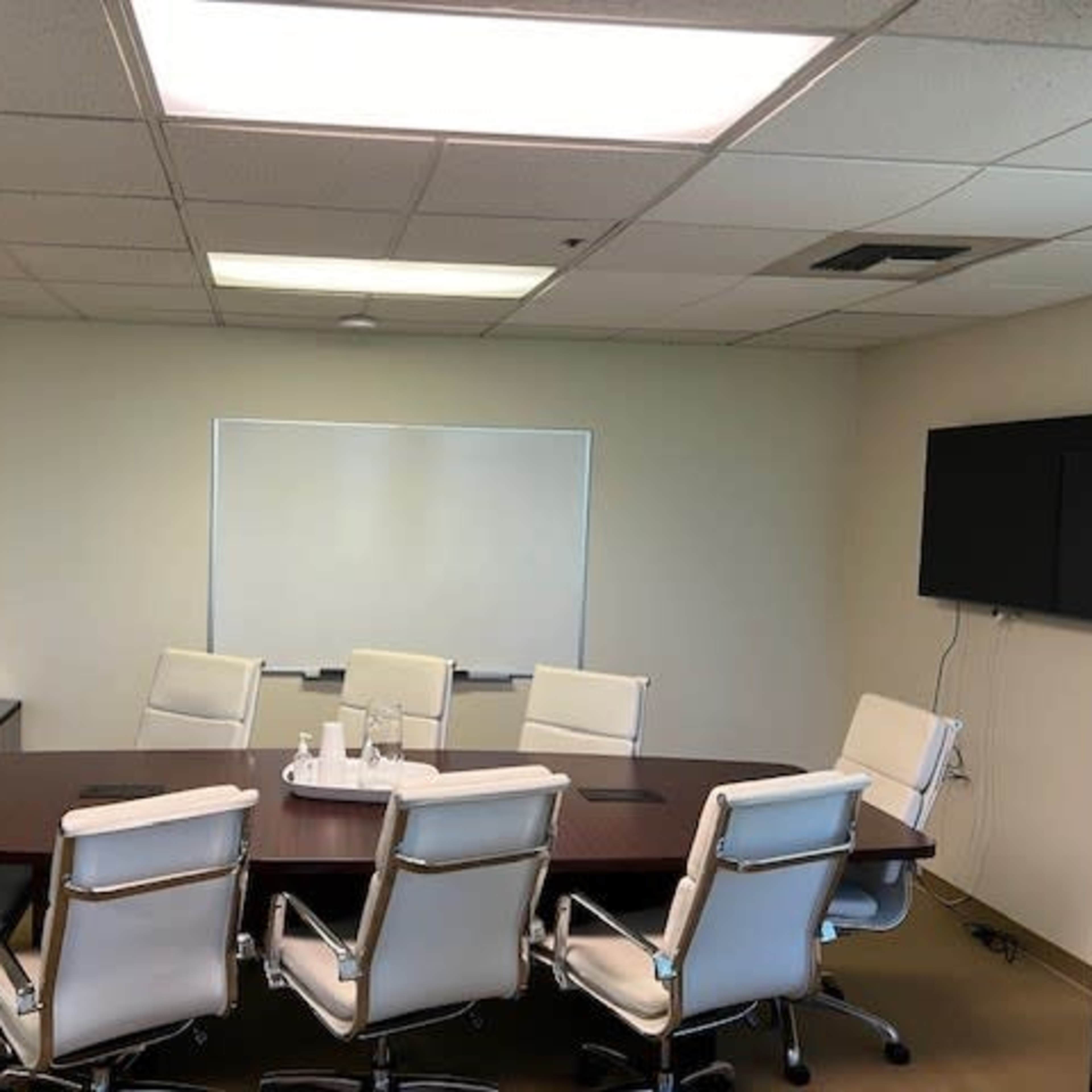 A conference room features a large rectangular table surrounded by eight white ergonomic chairs, a whiteboard on one wall, and a wall-mounted television on another.