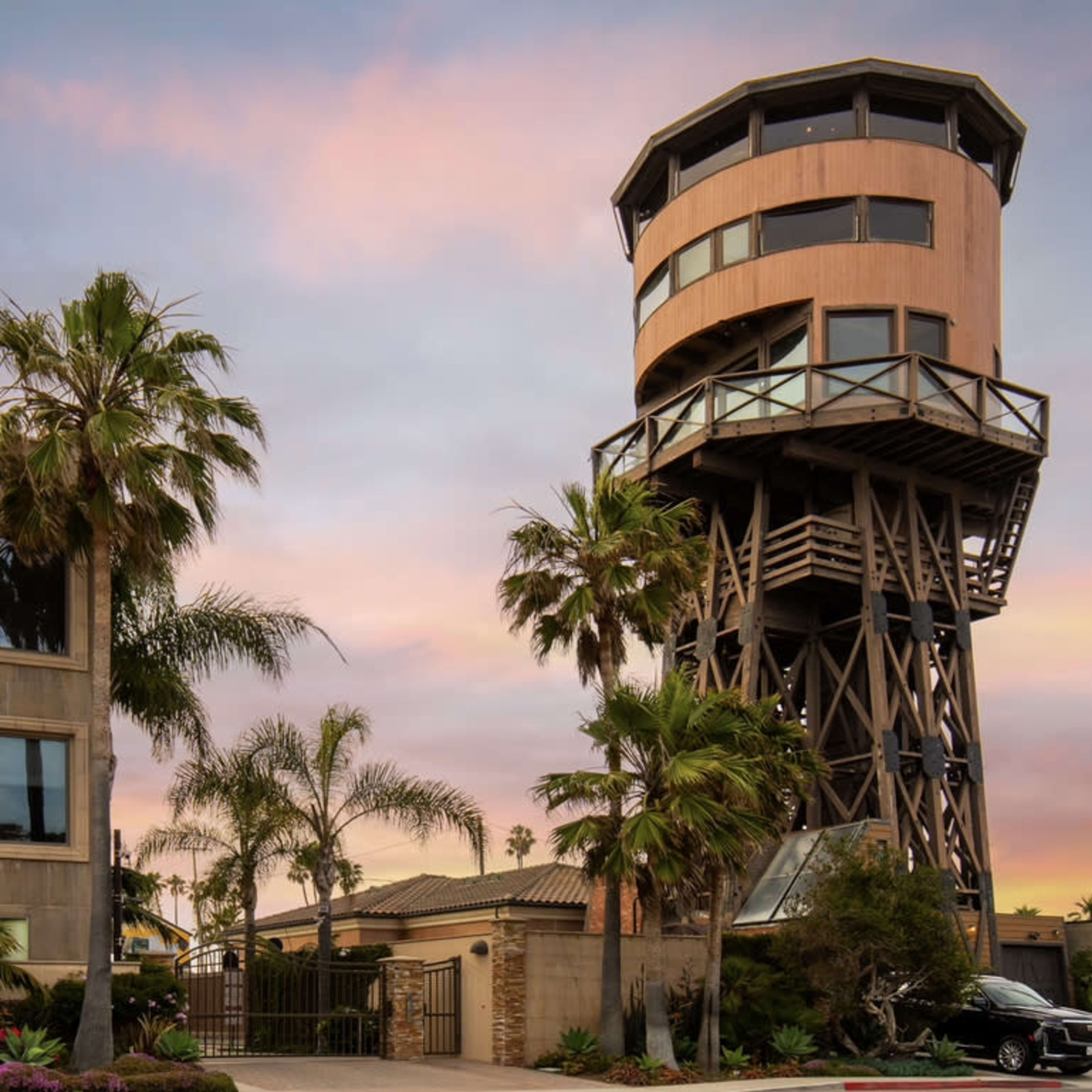 A tall observation tower made of wood and metal, surrounded by palm trees and a modern building, set against a colorful sunset sky.