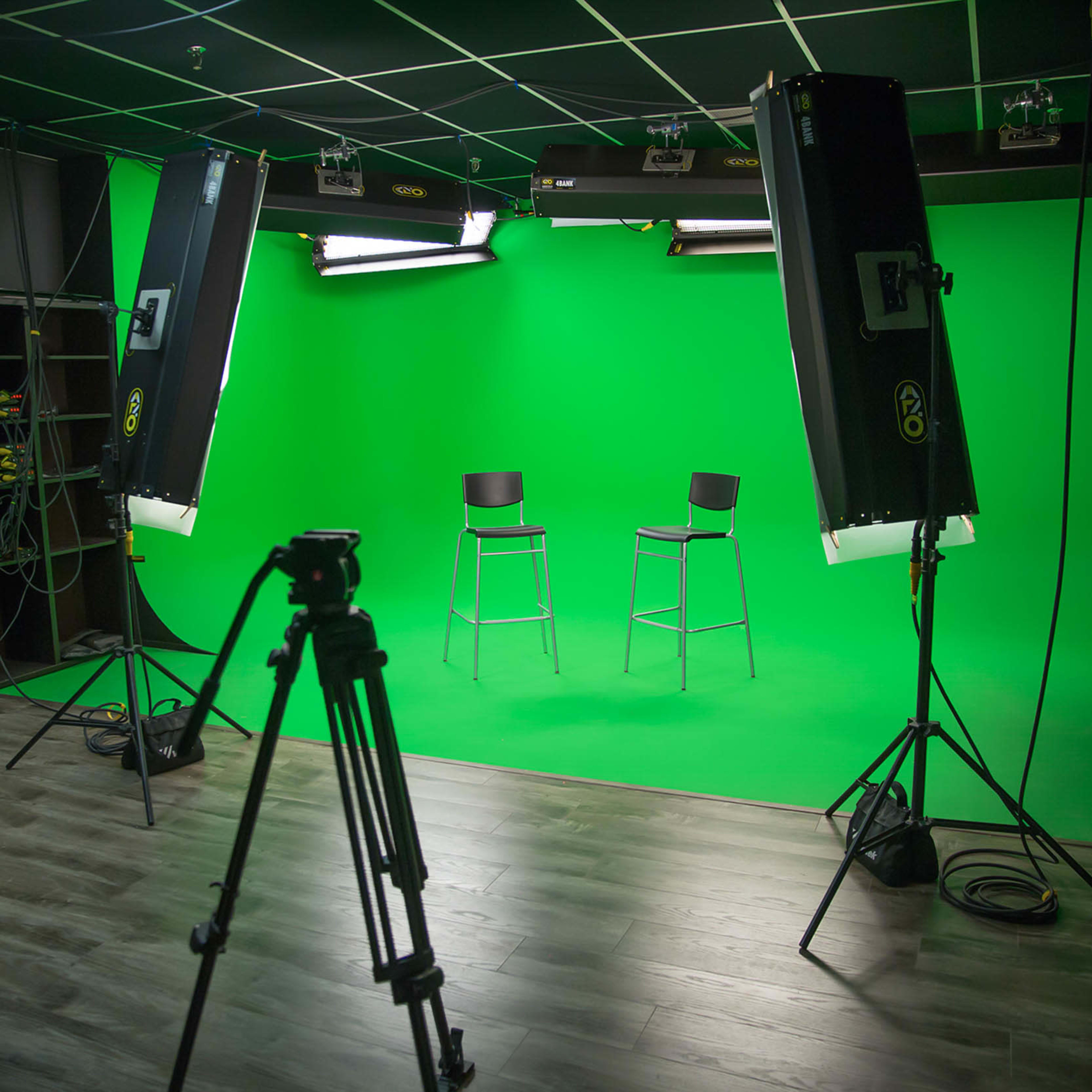 A video production studio with two high stools positioned in front of a green screen, flanked by large lights and a tripod.