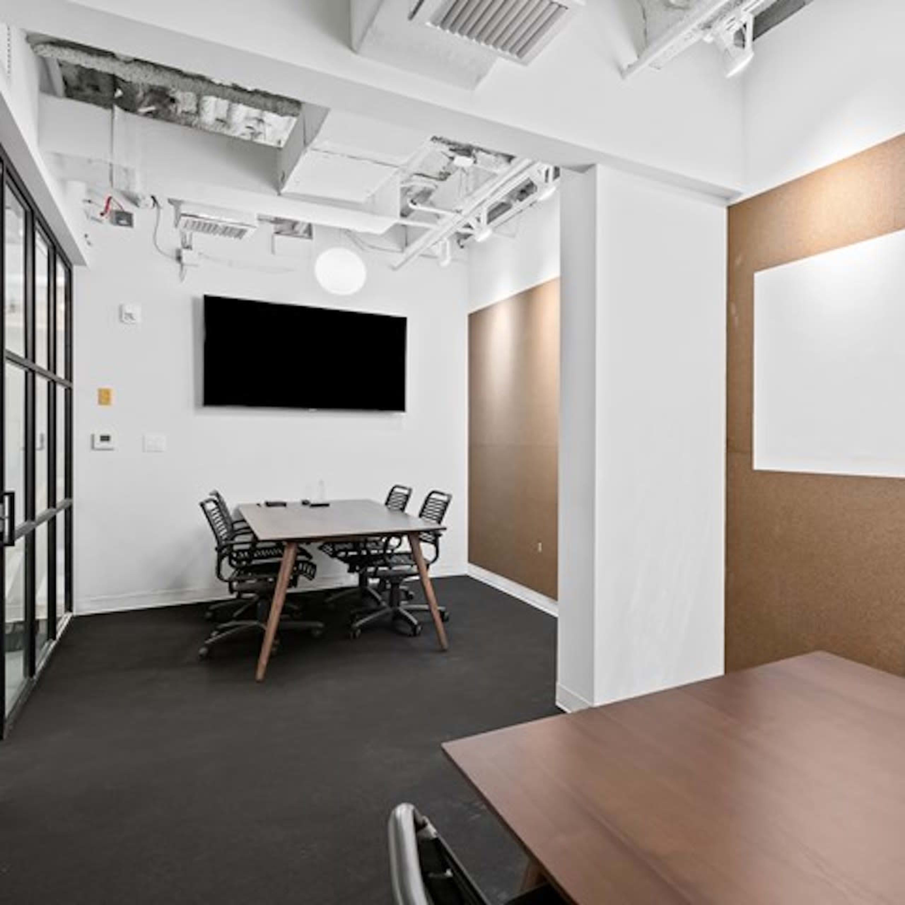 A conference room features a long wooden table, black chairs, and cork walls, with a television mounted on one side.