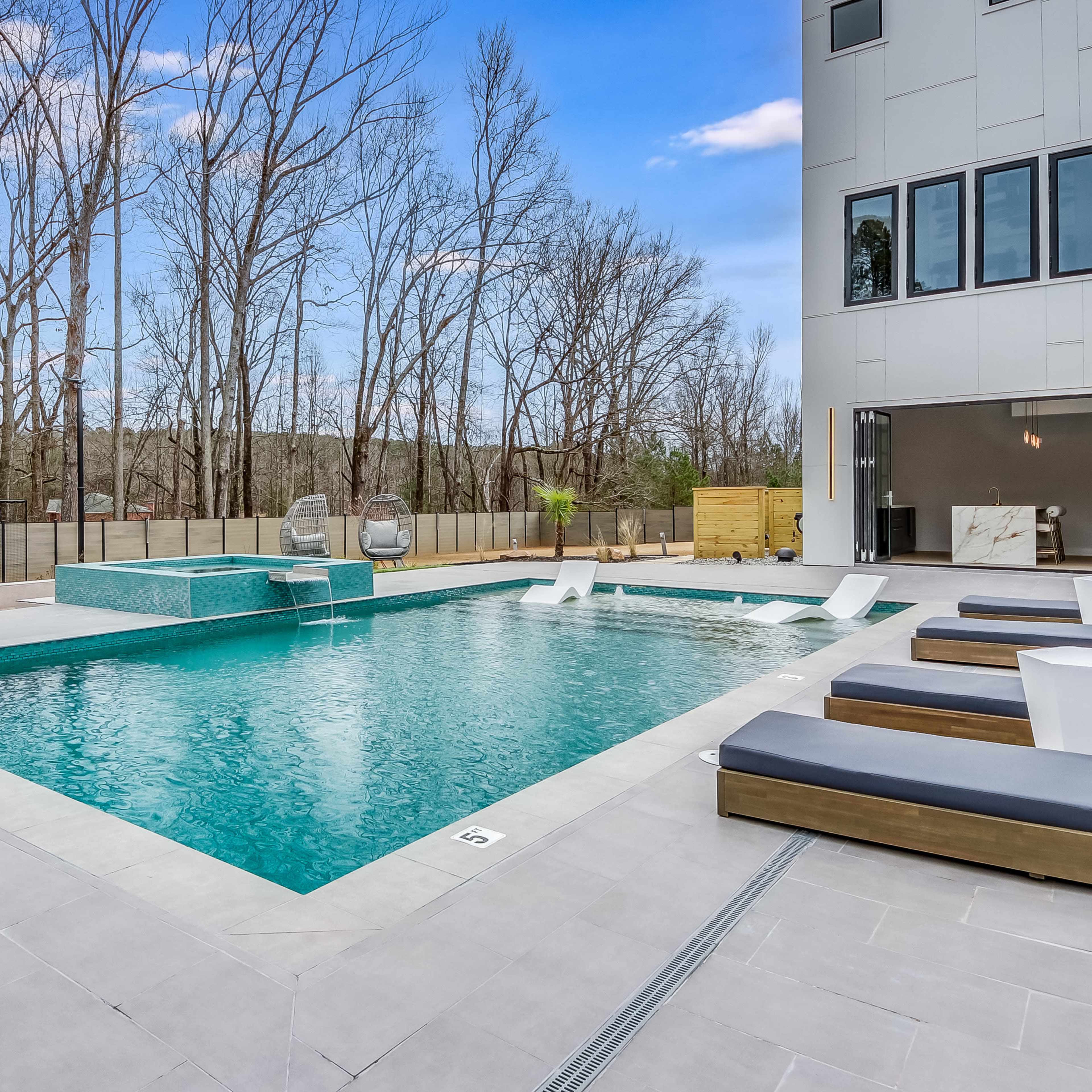 A modern outdoor pool area featuring a rectangular pool with lounge chairs and a basketball hoop in the background, surrounded by bare trees.