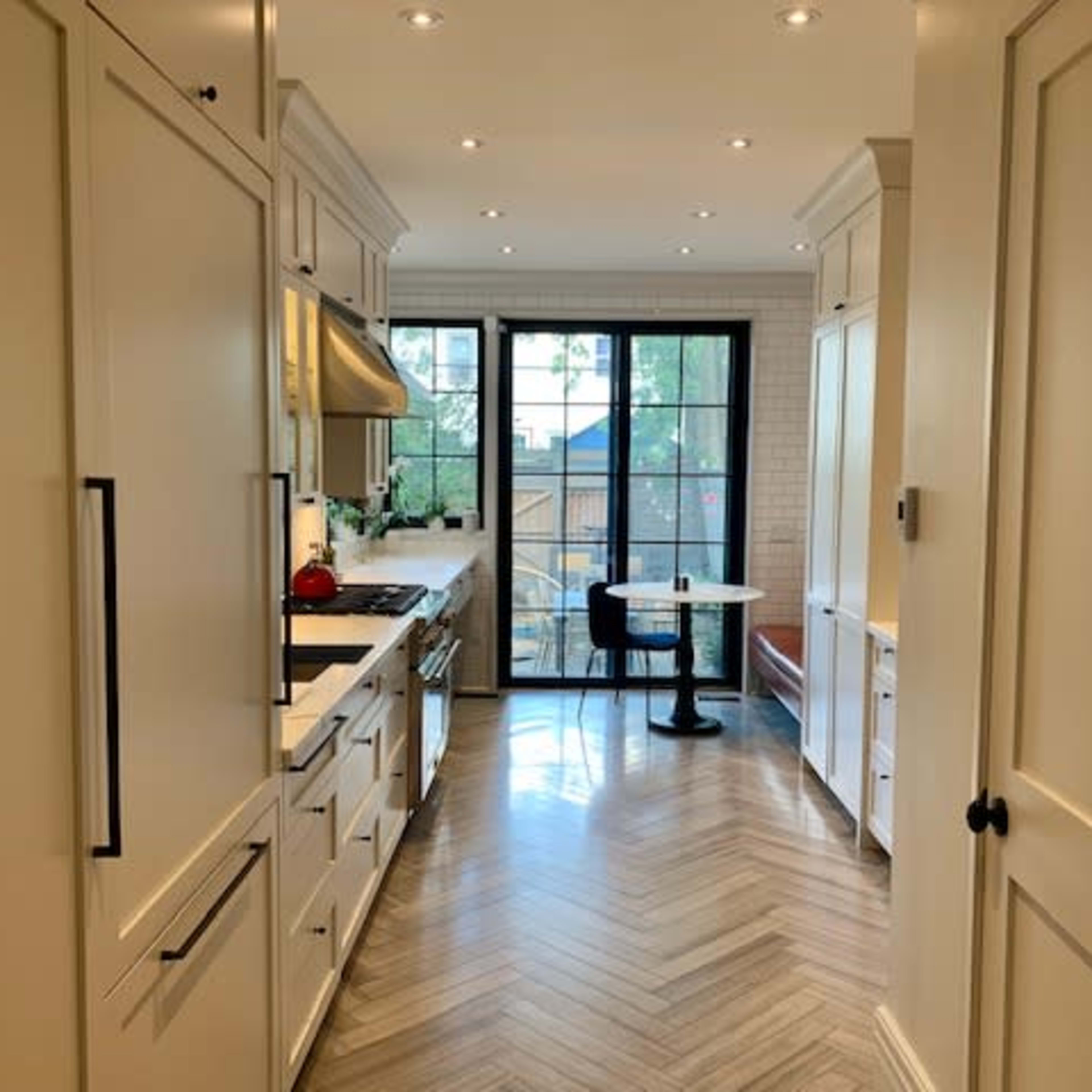 A modern kitchen features light cabinets, a herringbone floor, and a dining table set near a large window.