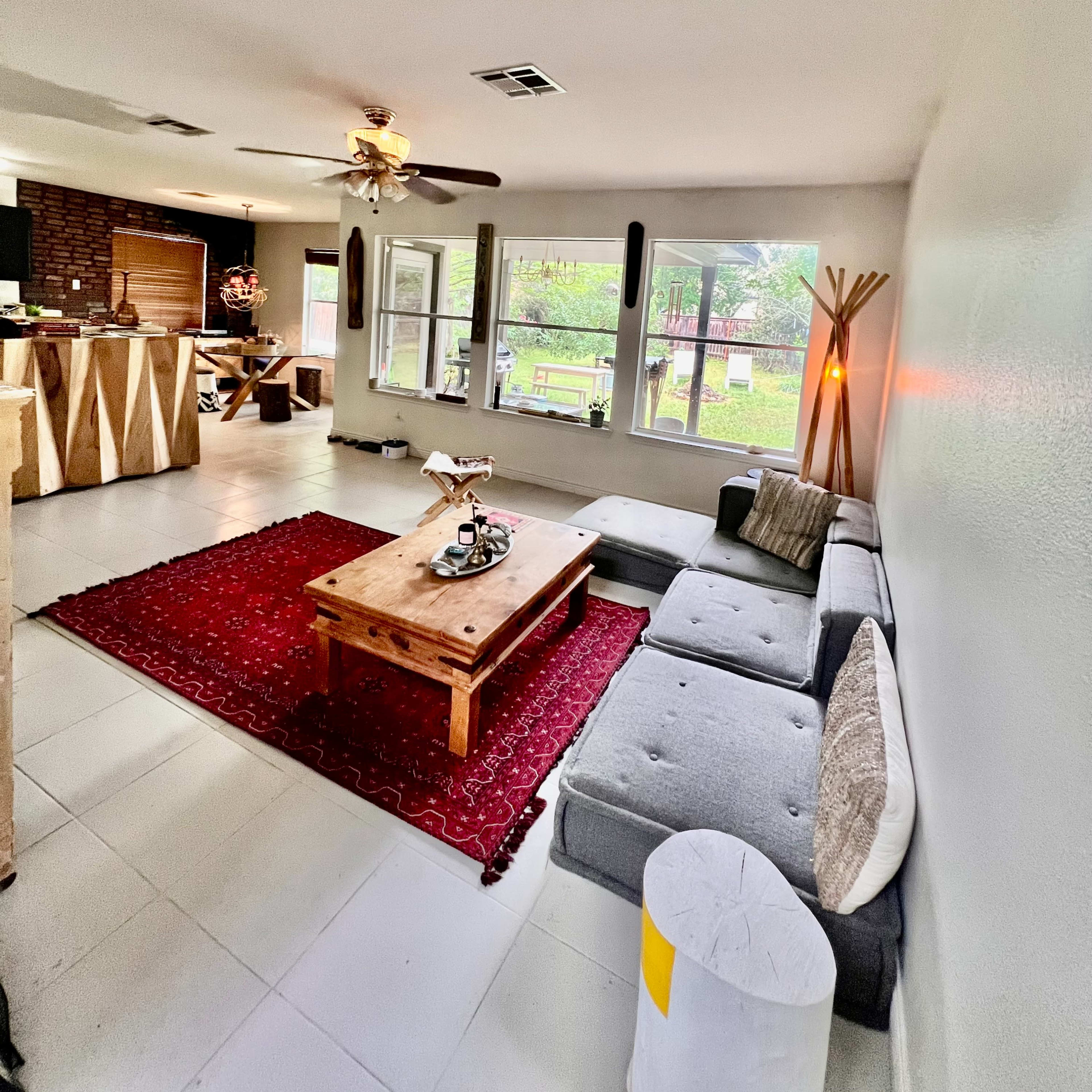 A living room with a gray sectional sofa, a wooden coffee table on a red area rug, and large windows providing natural light.