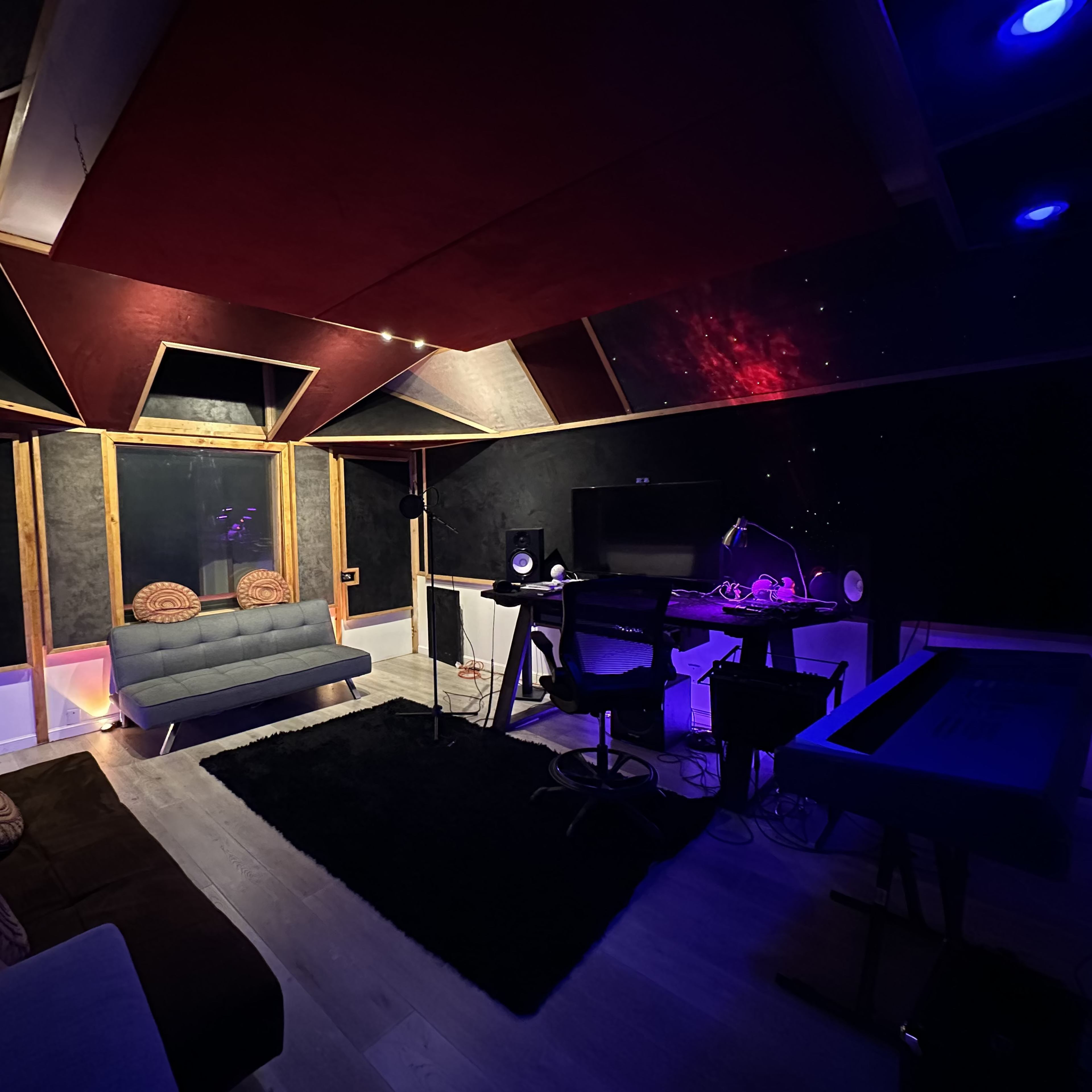 A music studio with a sectional couch, a desk with a computer and audio equipment, and colorful lighting on the ceiling.