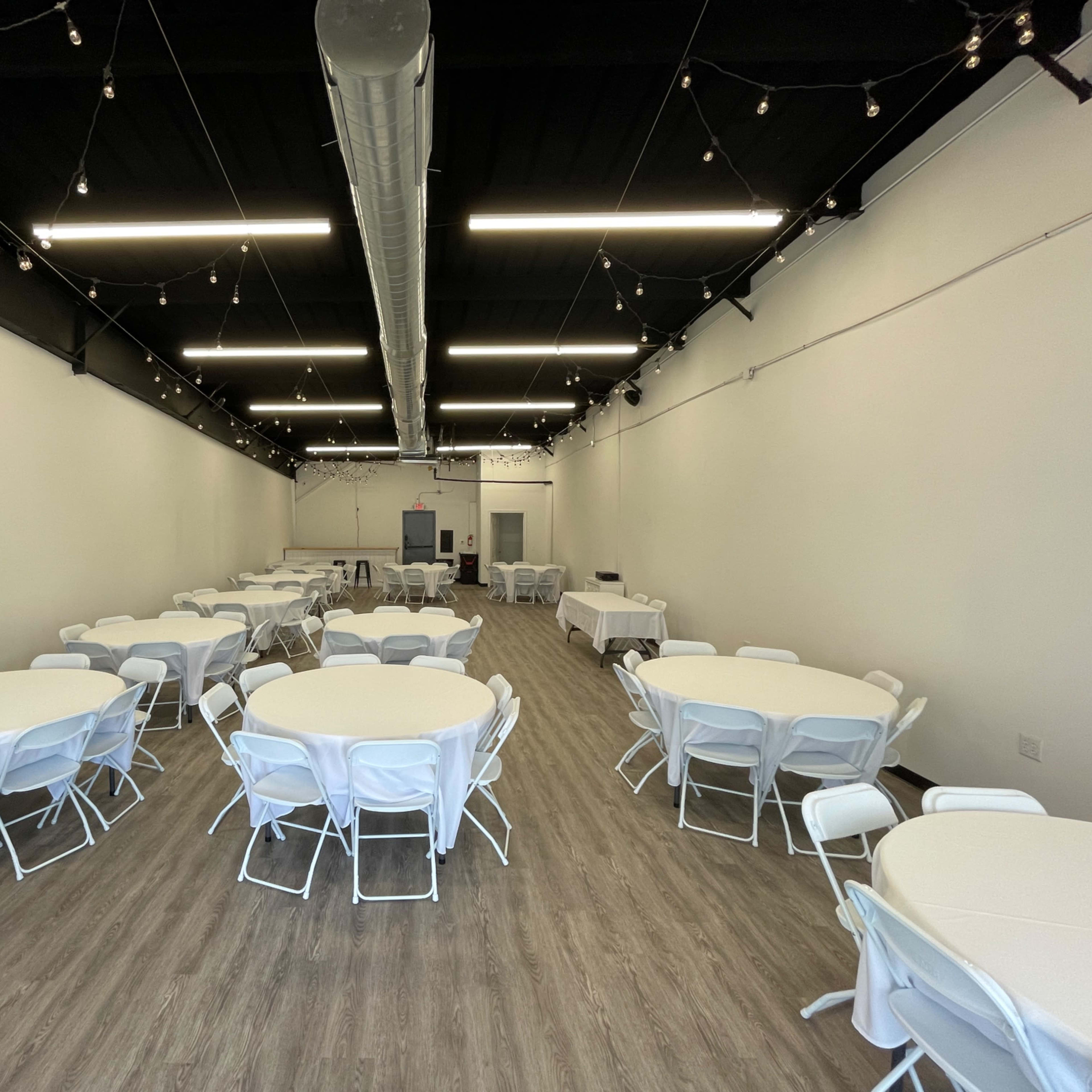 A large, well-lit event space with several round tables covered in white tablecloths and arranged neatly across the floor.