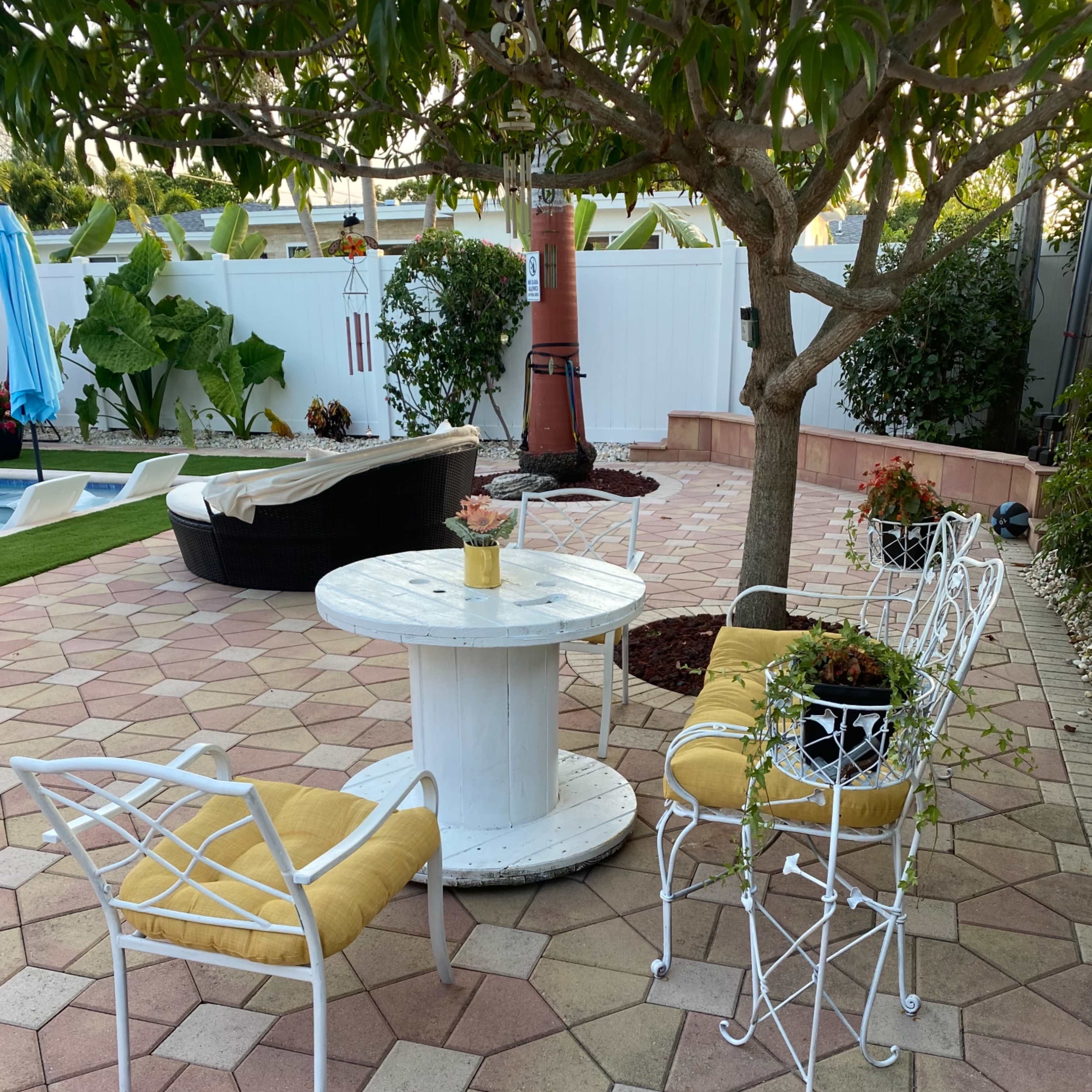 A circular white table surrounded by four light-colored chairs sits on a patterned stone patio under a tree, with a lush garden in the background.