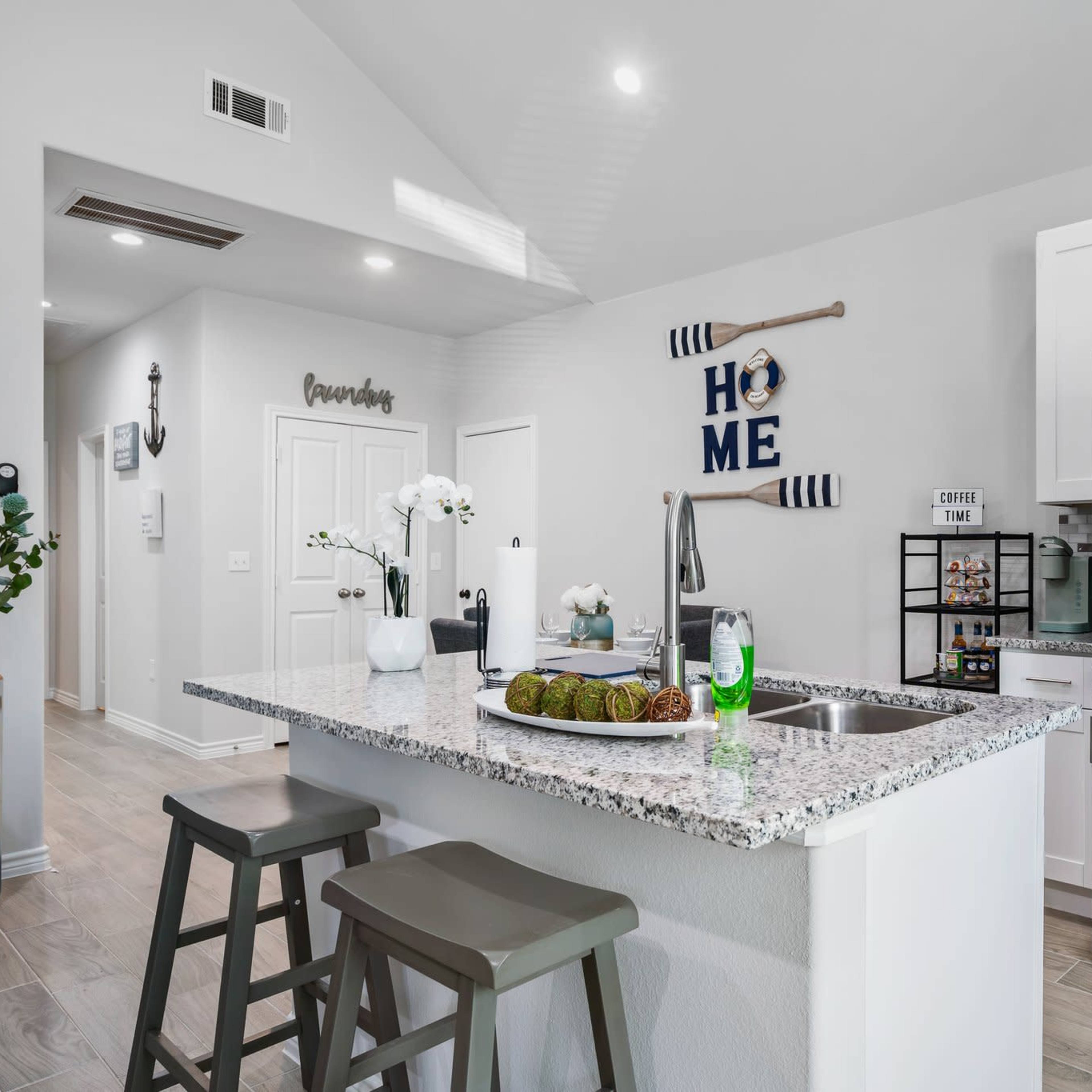 A modern kitchen features a gray granite island with stools, stainless steel appliances, and decorative items on the countertop.