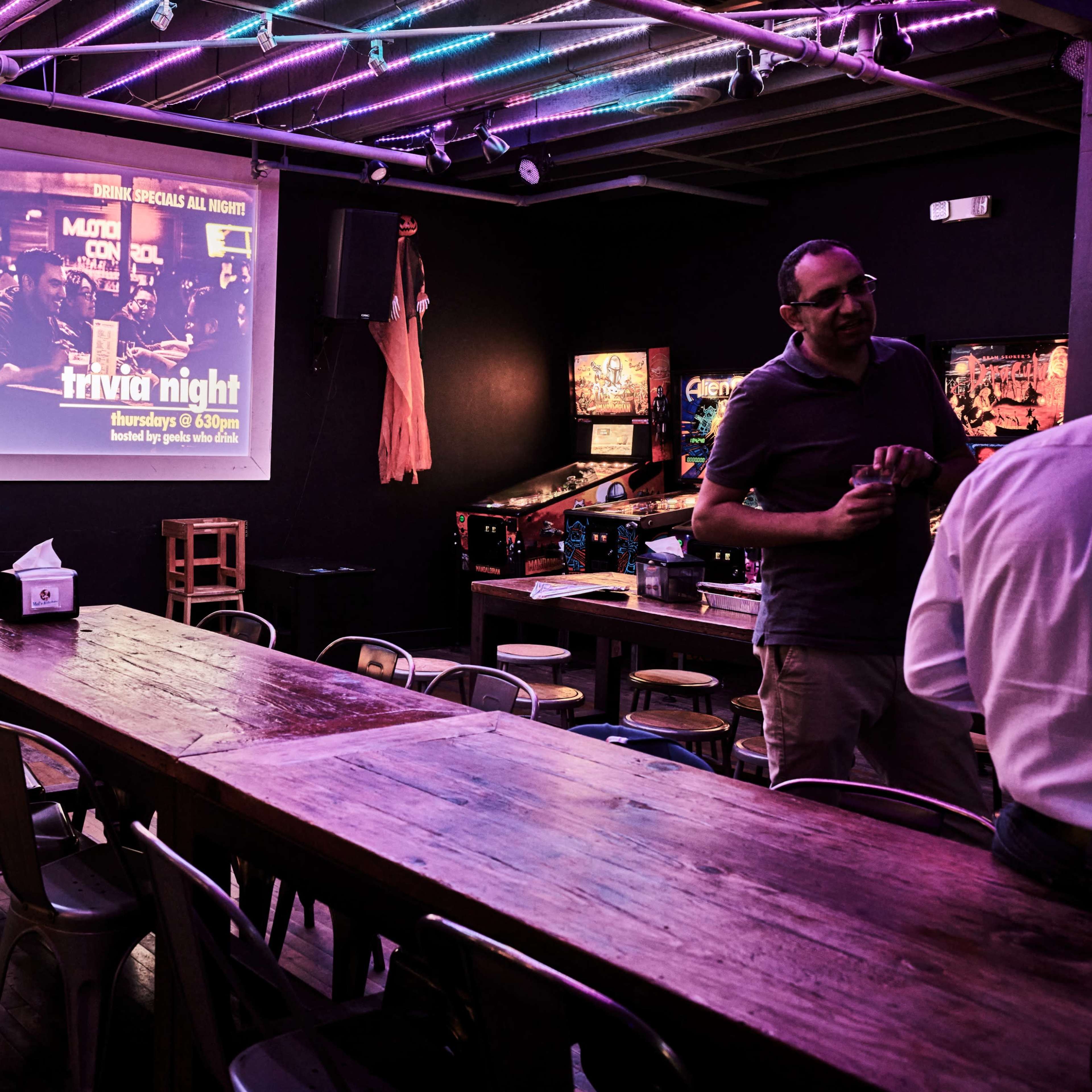 A dimly lit bar with a long wooden table and a projector screen displaying a trivia night advertisement on the wall.