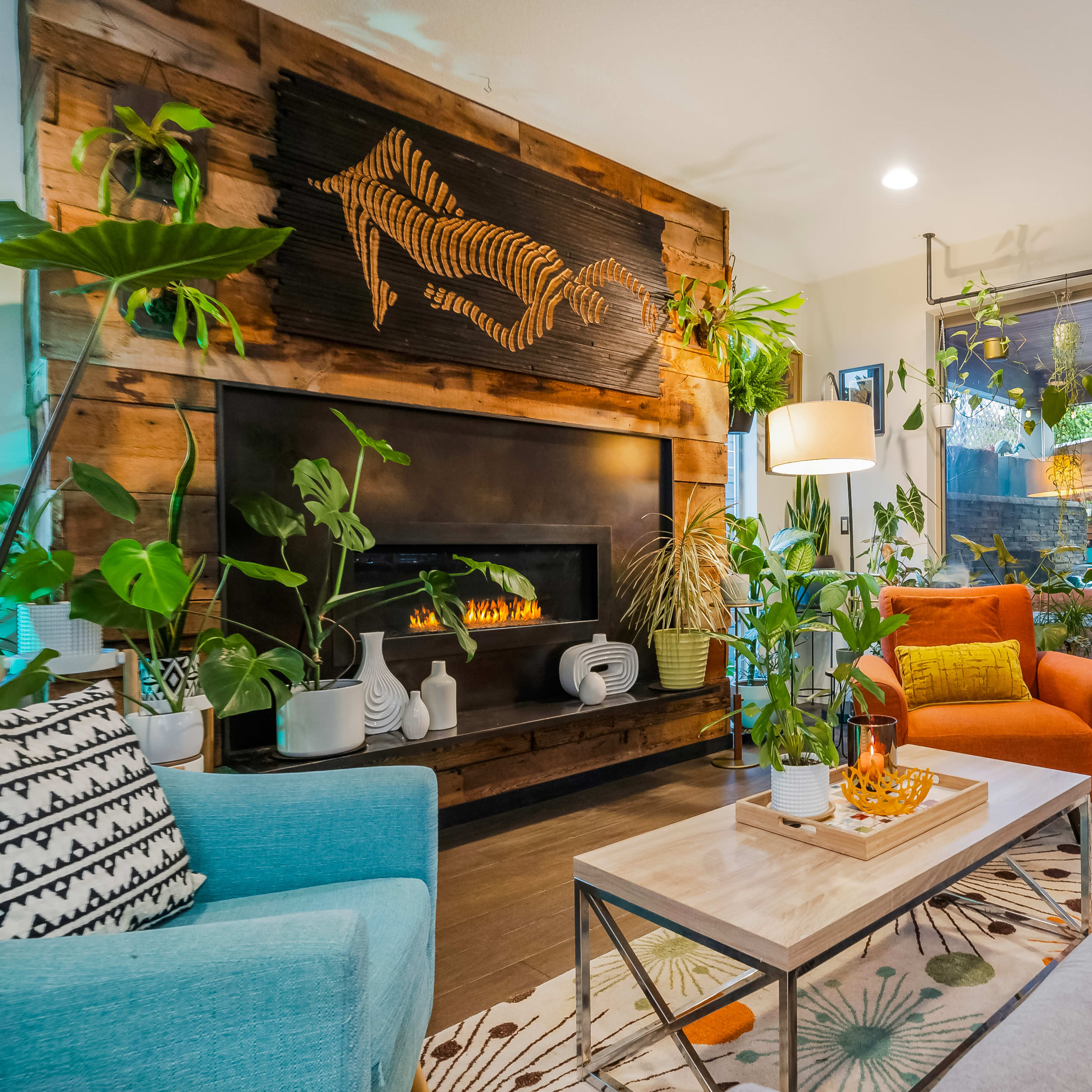 A modern living room with a wooden fireplace adorned with a decorative wall piece, colorful seating, and various indoor plants.