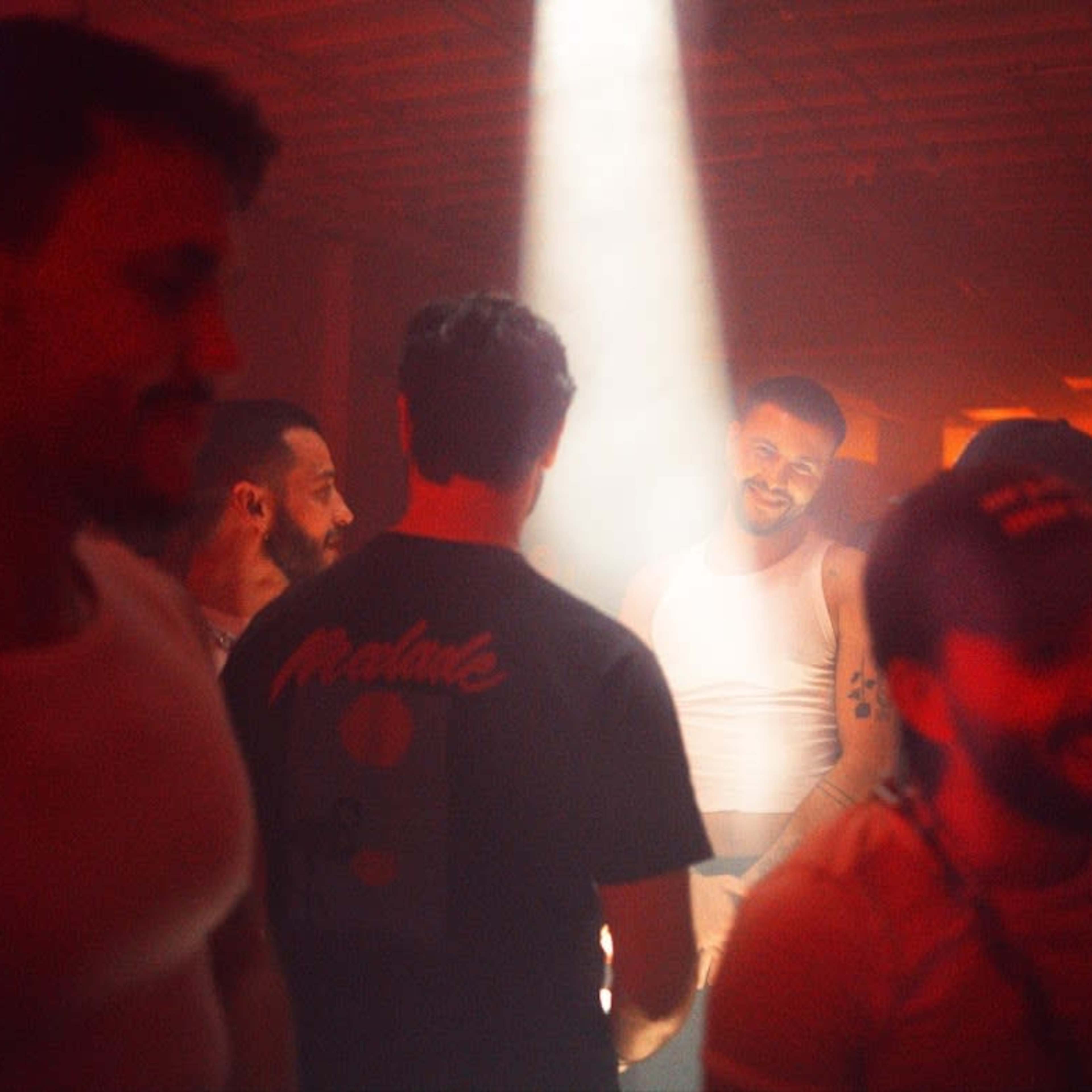 A group of men is gathered in a dimly lit space, with a beam of light illuminating one individual in a white tank top.