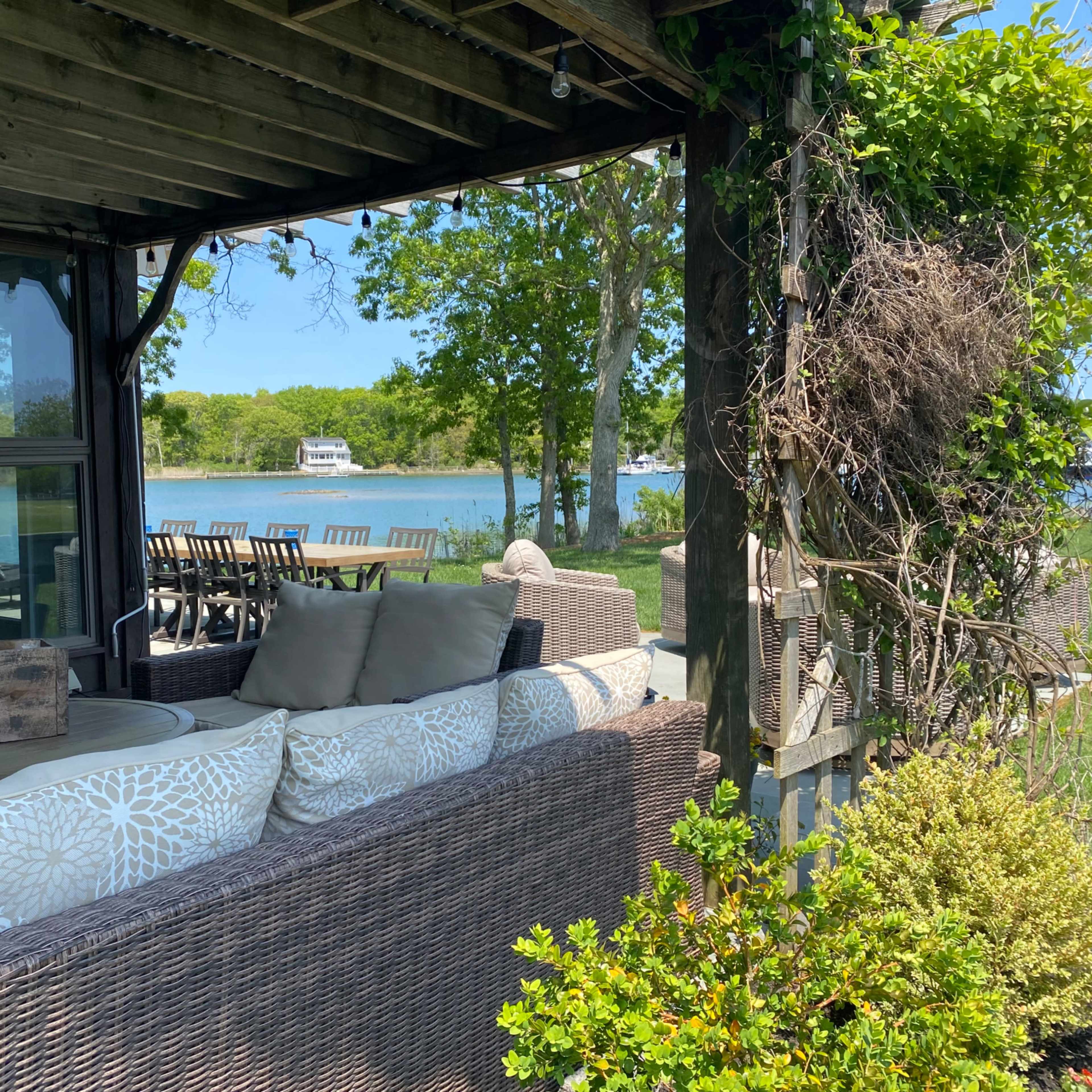 A covered patio with wicker furniture overlooks a calm waterfront with boats in the distance.