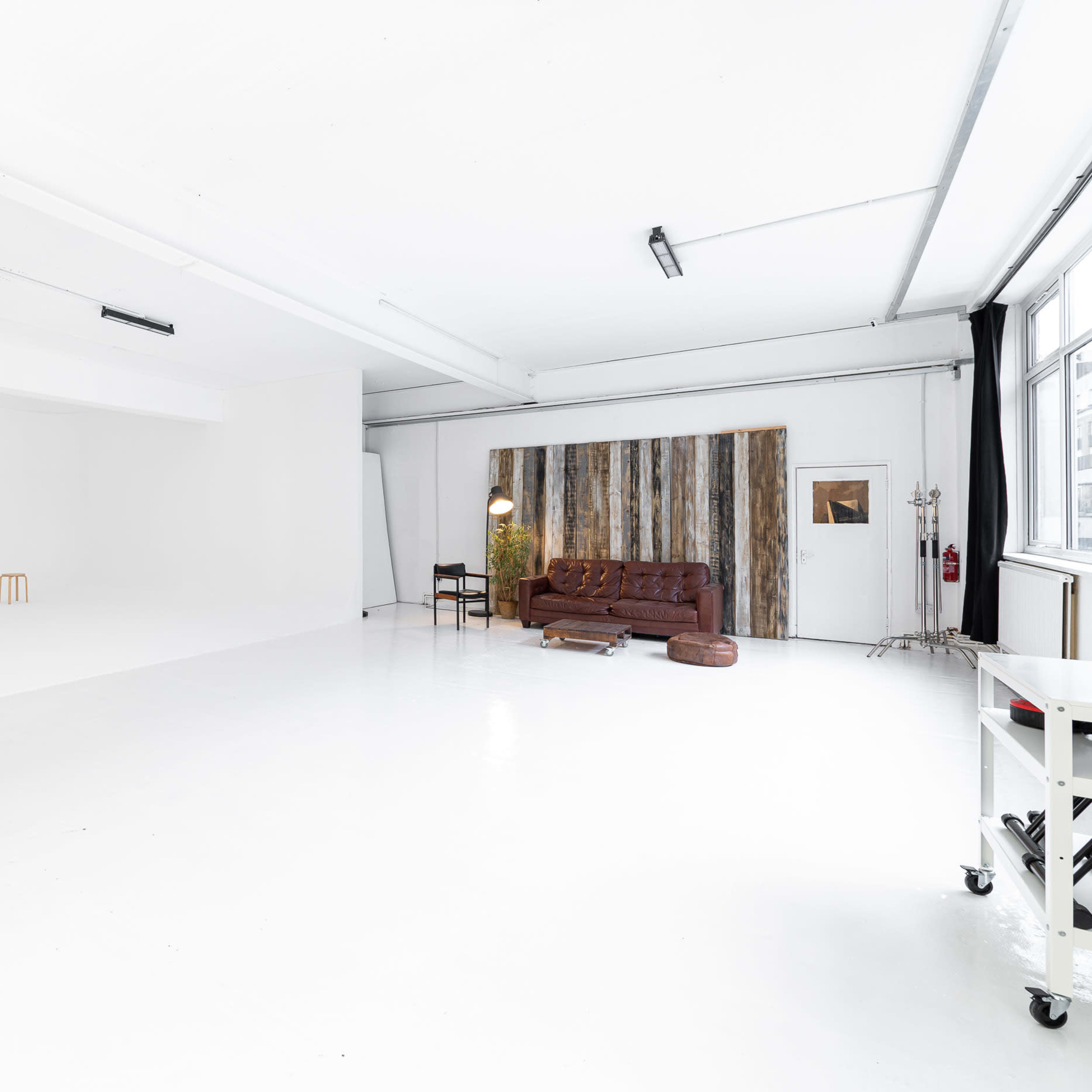 A large, empty studio space with white walls and floors, featuring a wooden accent wall, a brown couch, and a standing lamp.