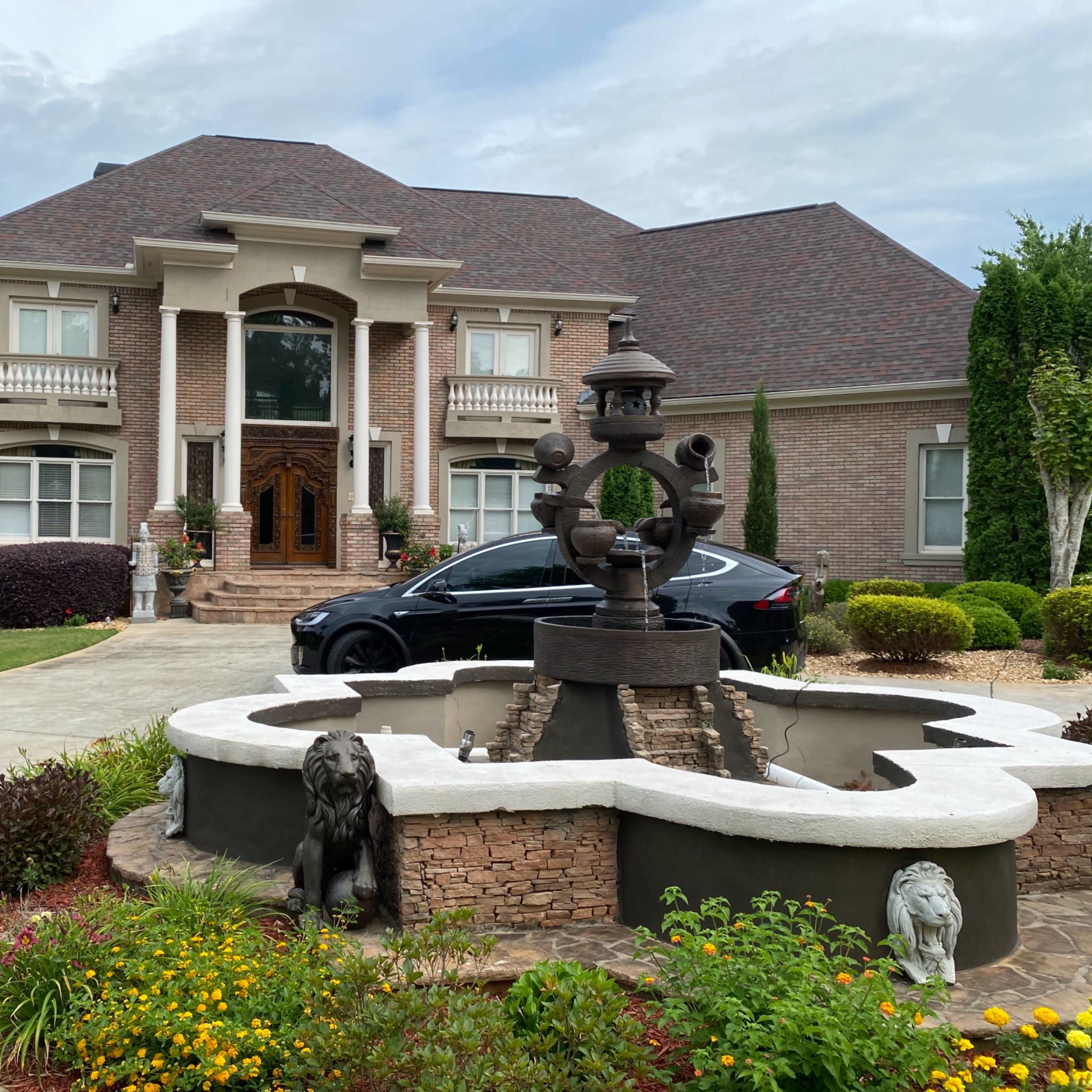 A large brick house with a fountain and sculptures in the front yard, alongside a driveway leading to a black car.
