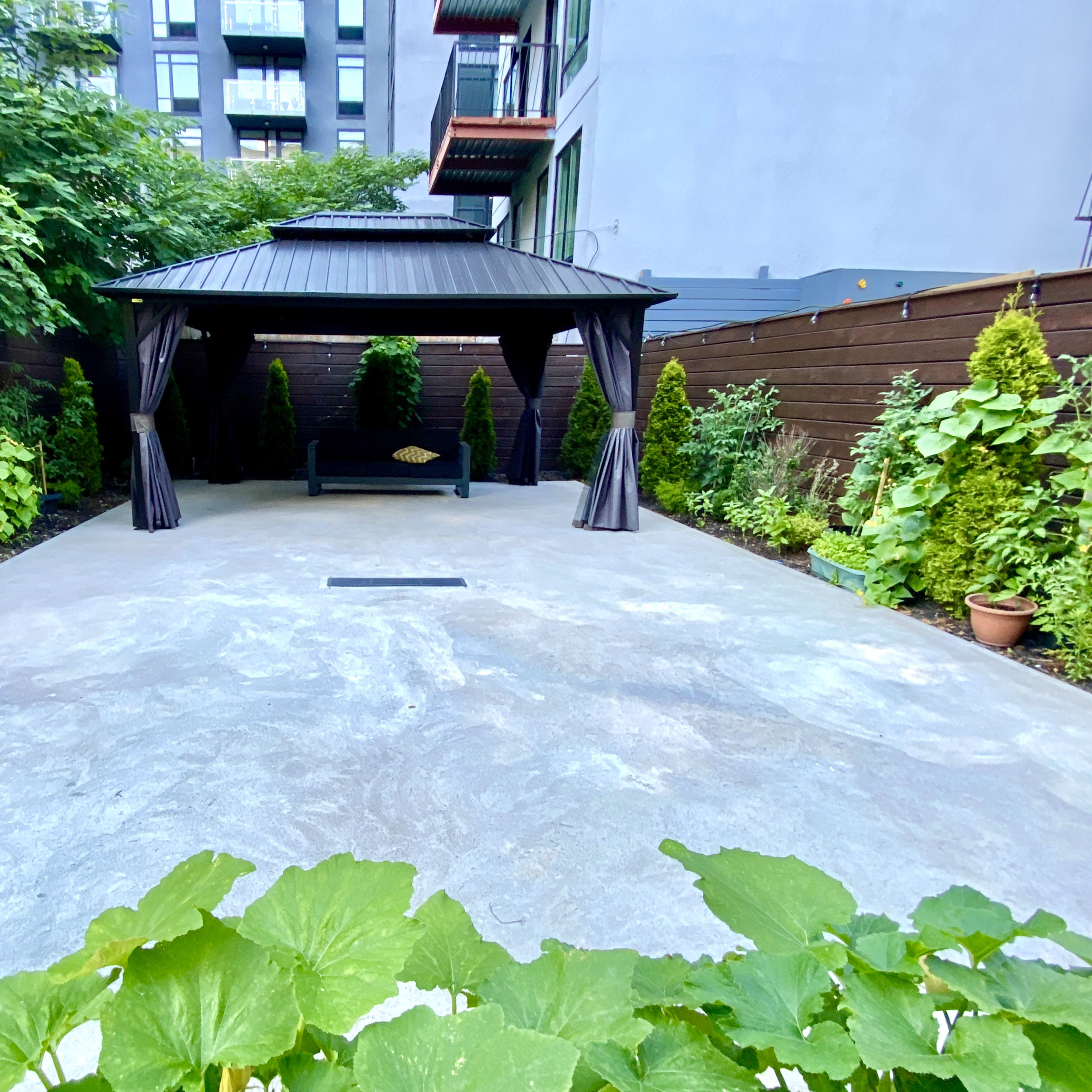 A concrete outdoor area featuring a gazebo surrounded by neatly arranged greenery, including small shrubs and potted plants.
