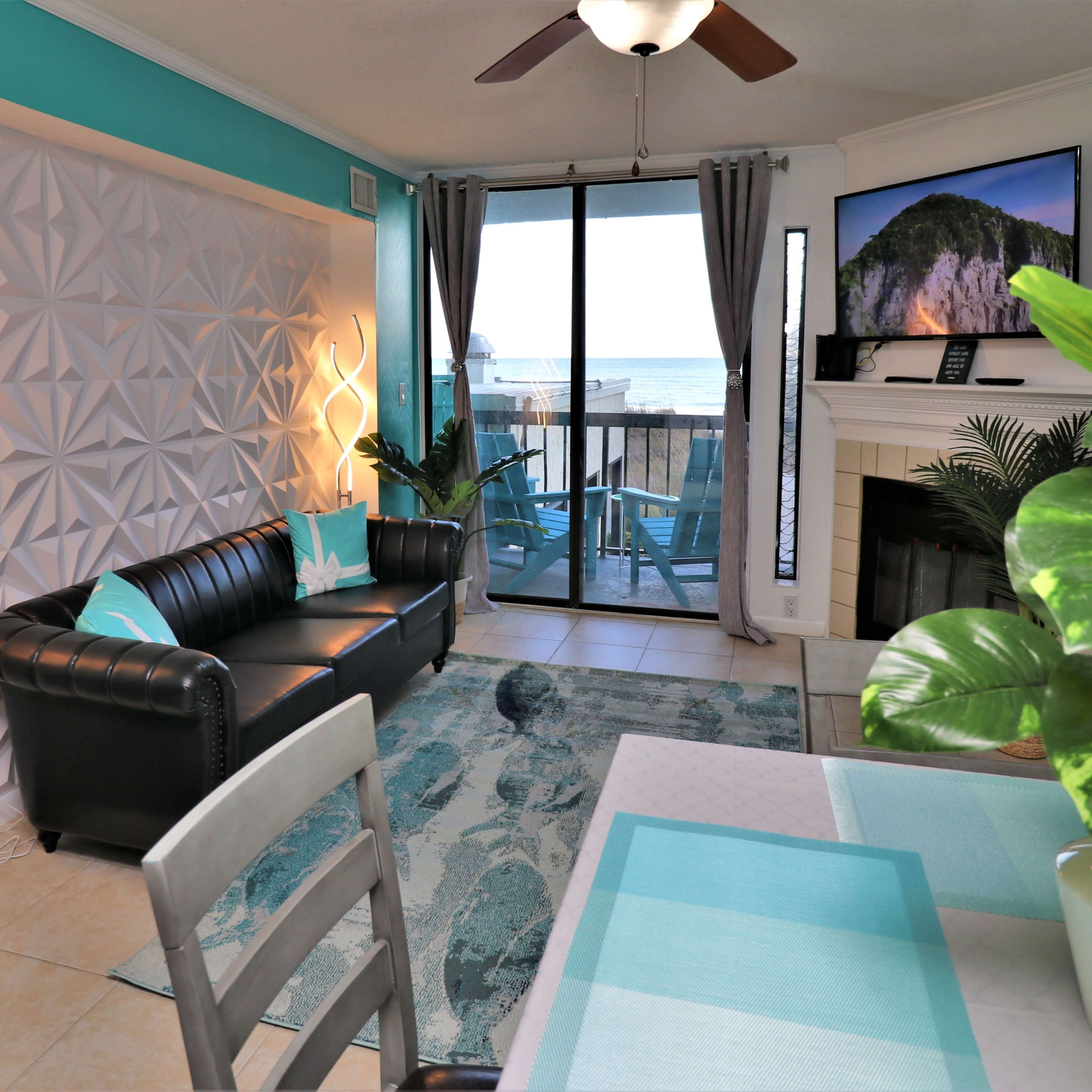 A modern living room with a black leather sofa, a patterned feature wall, and a large window overlooking the ocean.