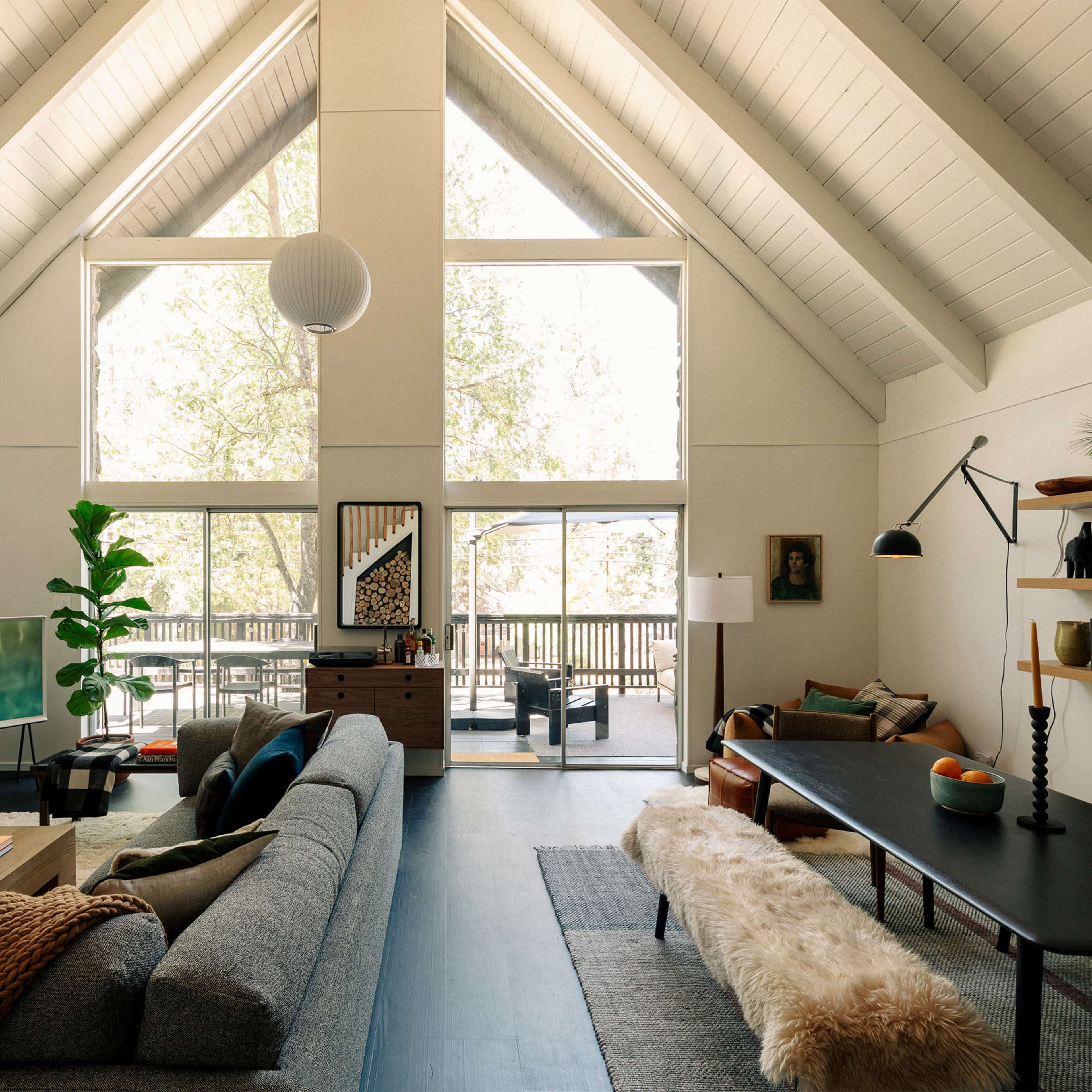 A spacious living room with high ceilings, large windows, a modern sofa, a coffee table, and a television, all complemented by a natural view outside.