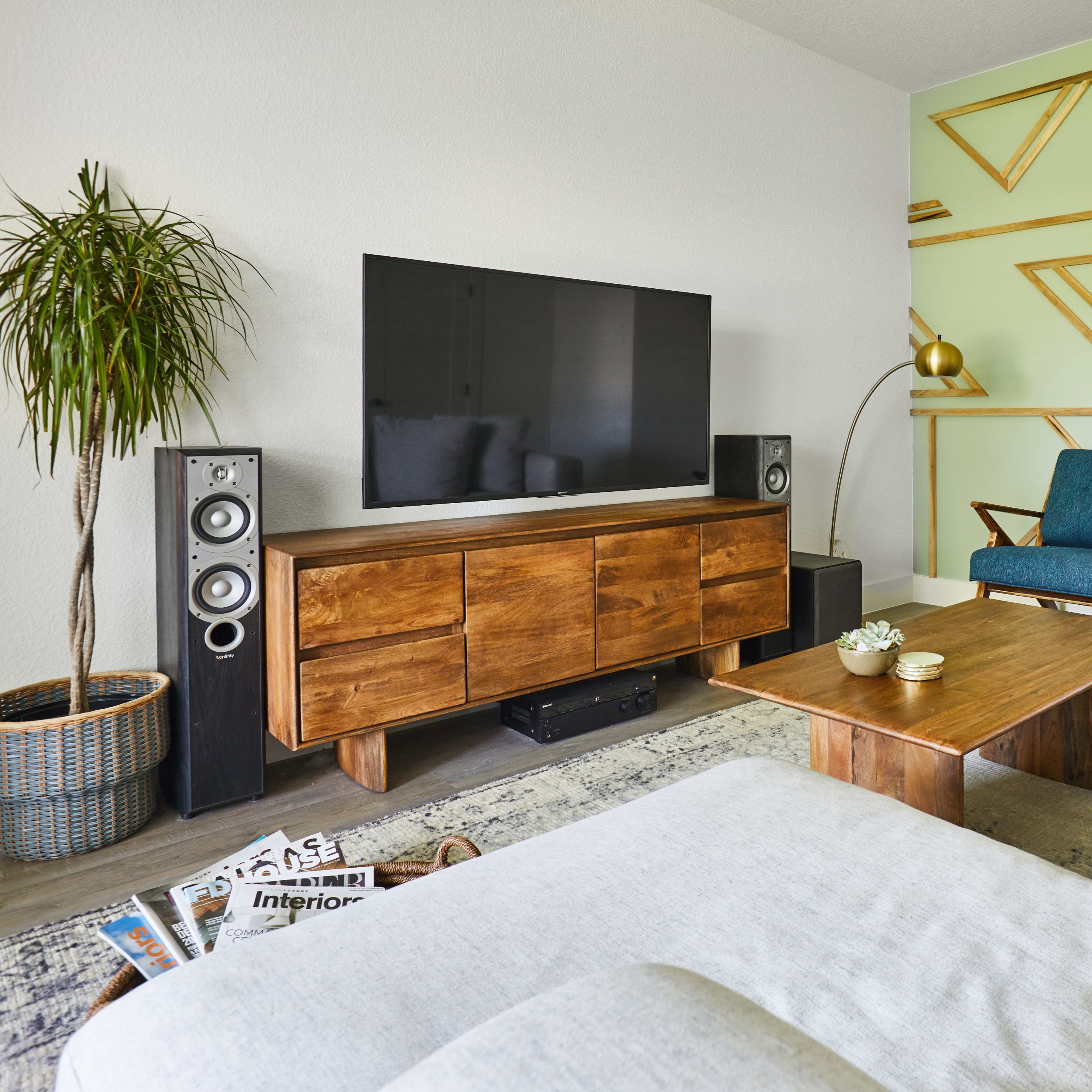 A modern living room features a light-colored sofa, a wooden coffee table, a TV mounted on the wall, and a chair next to a decorative plant.