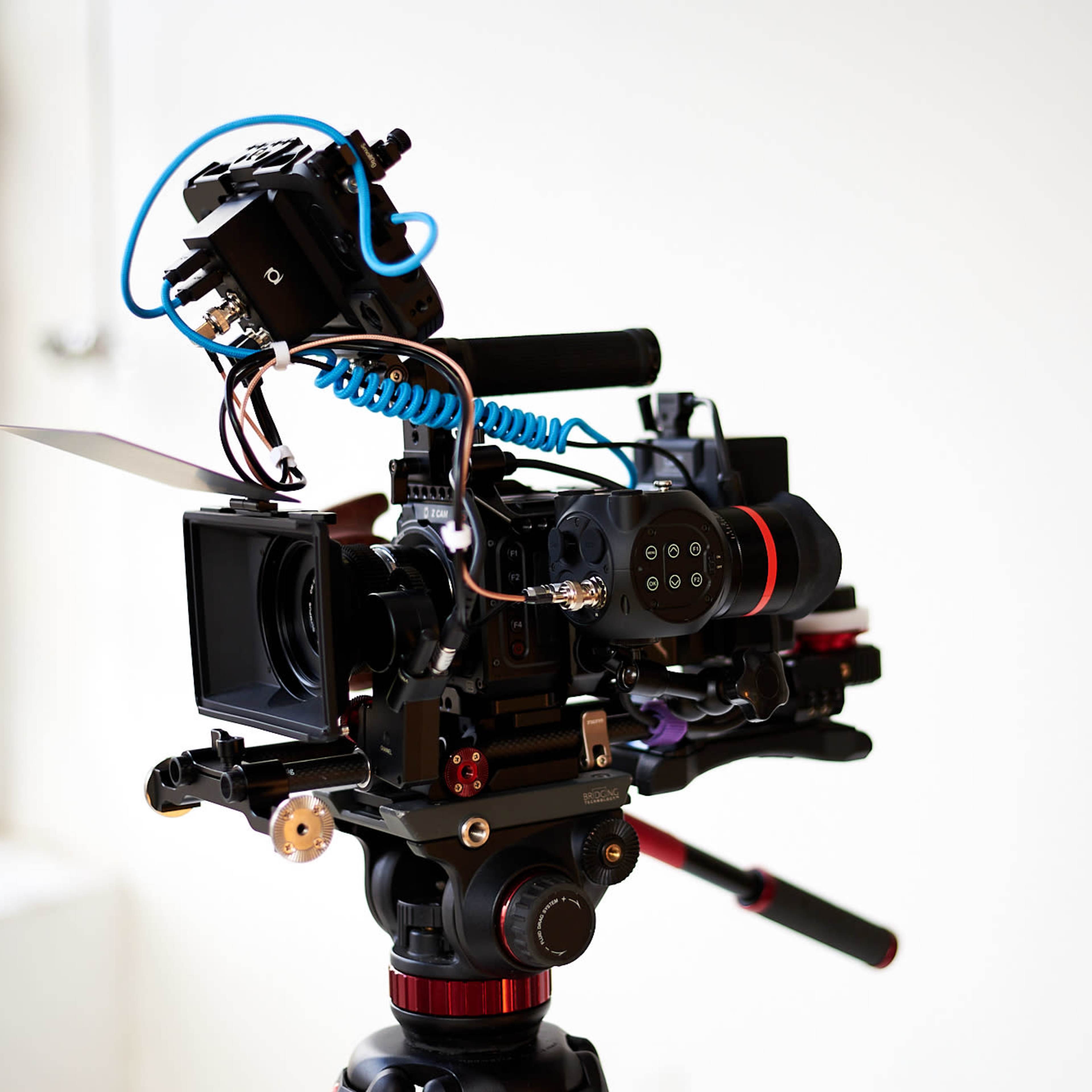 A professional video camera mounted on a tripod is positioned against a plain wall.