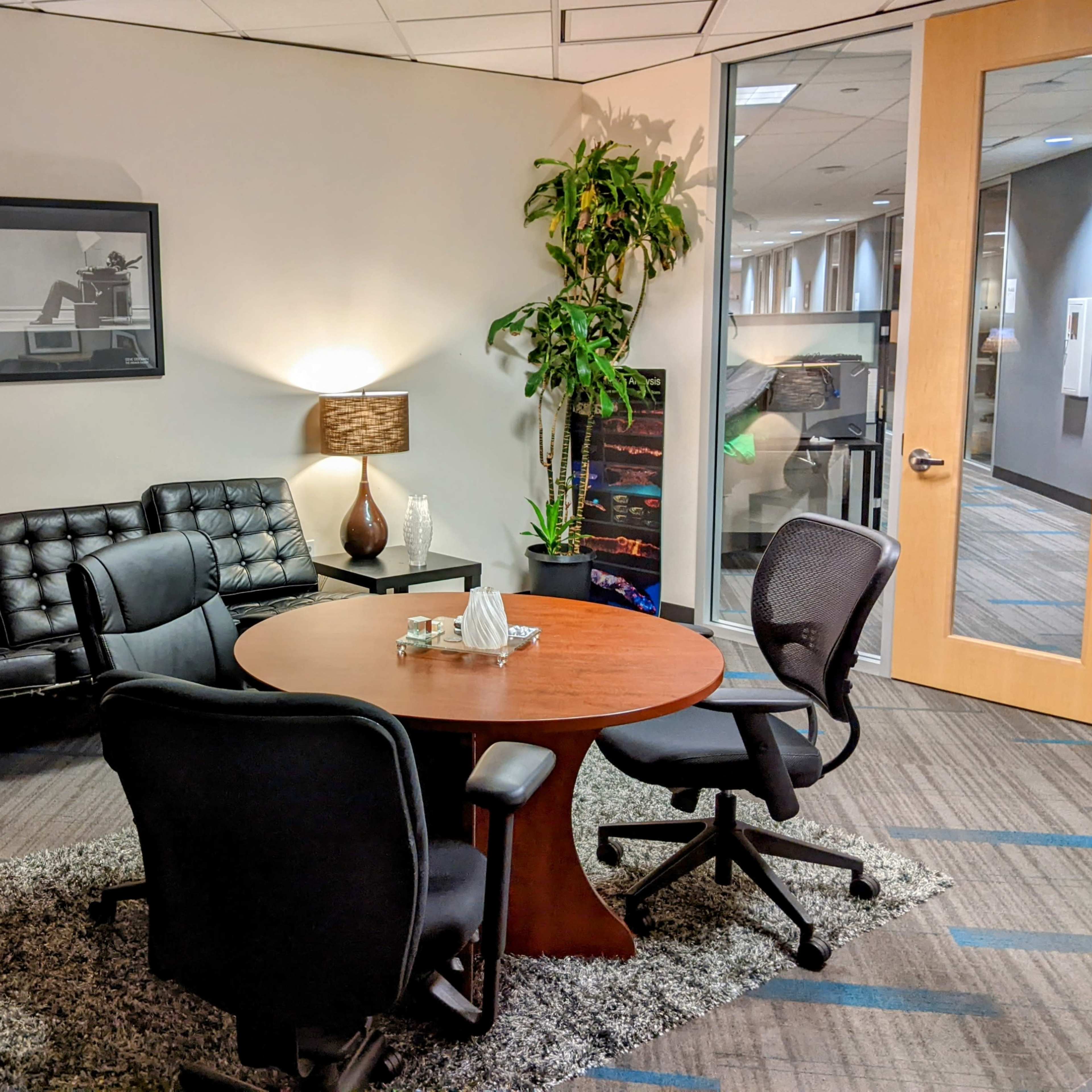 A modern office space features a round wooden table surrounded by chairs, a black leather couch, a lamp, and a potted plant.