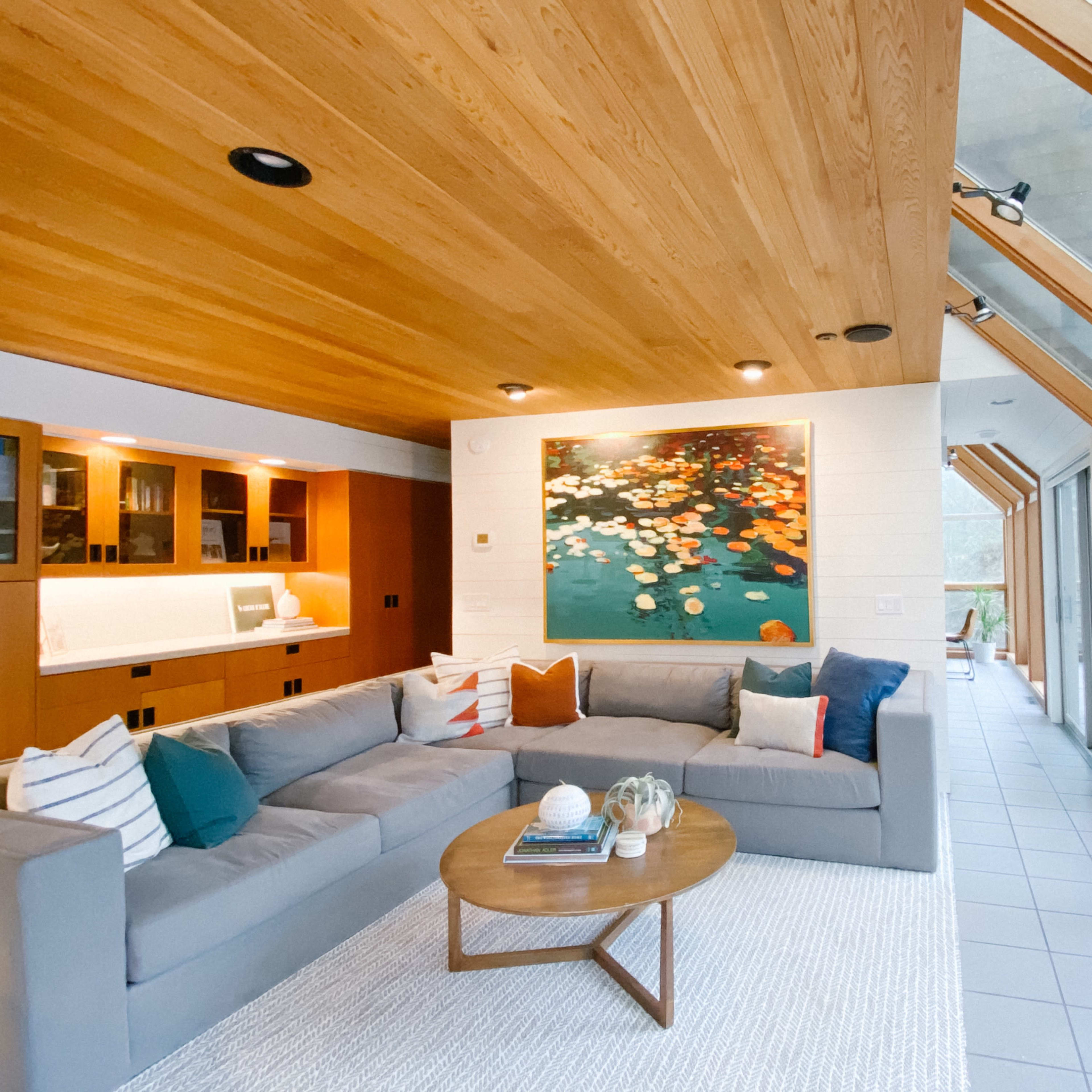 A spacious living room features a sectional sofa, a coffee table, and a large painting, with floor-to-ceiling windows that provide a view of the outdoors.