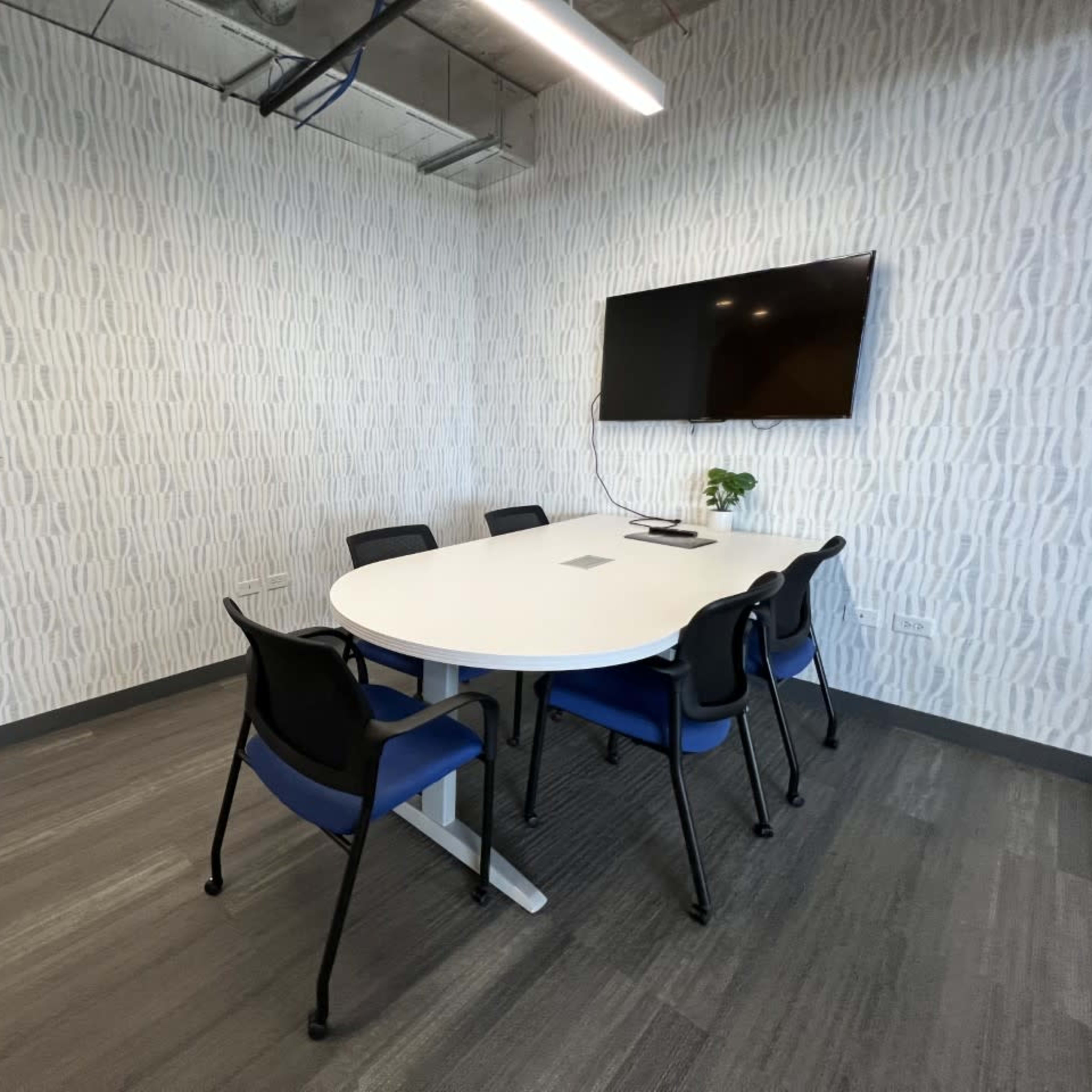 A modern conference room features a round table surrounded by six black and blue chairs, with a wall-mounted television and light-colored patterned wallpaper.