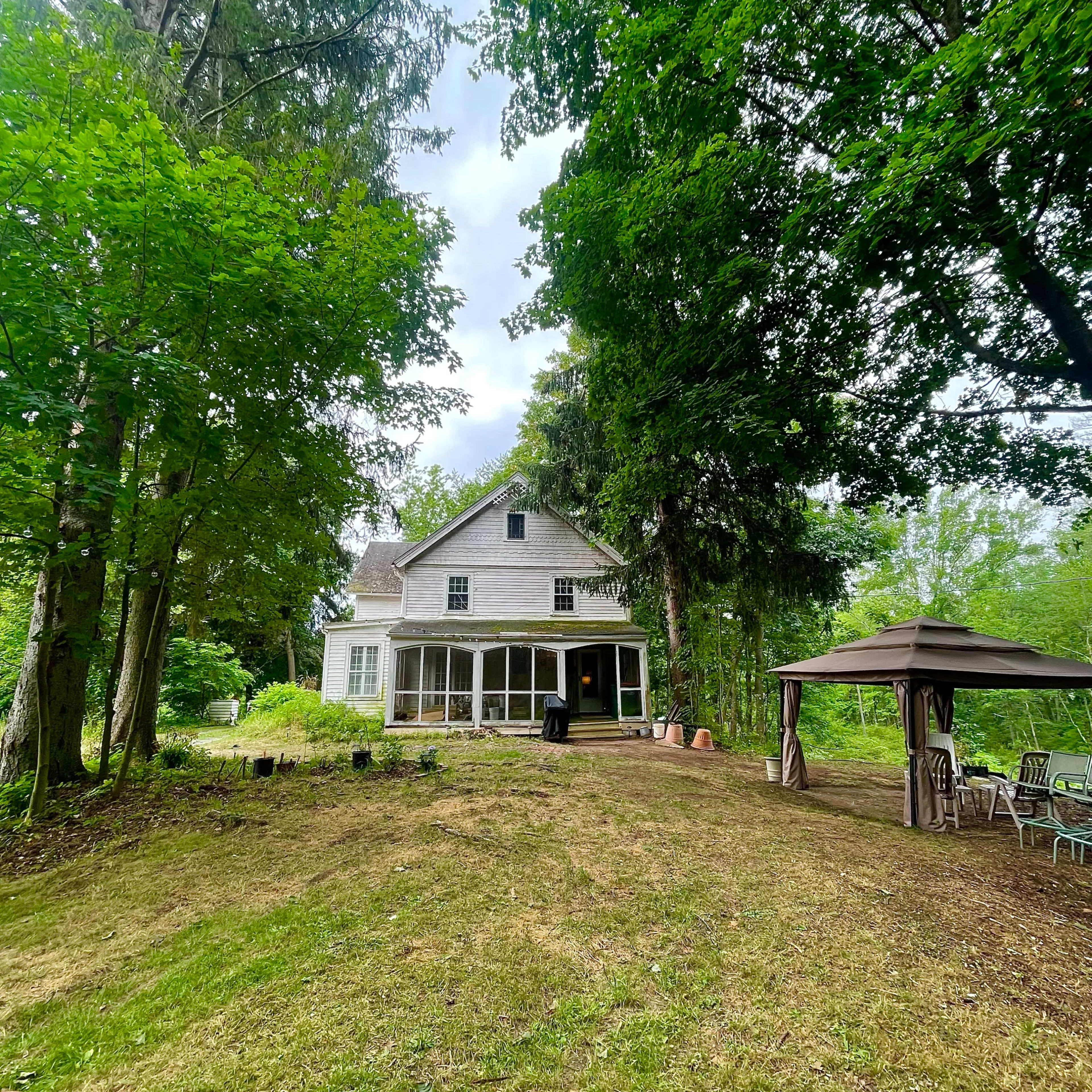 A two-story, weathered house is situated on a grassy slope, surrounded by tall trees and a gazebo nearby.