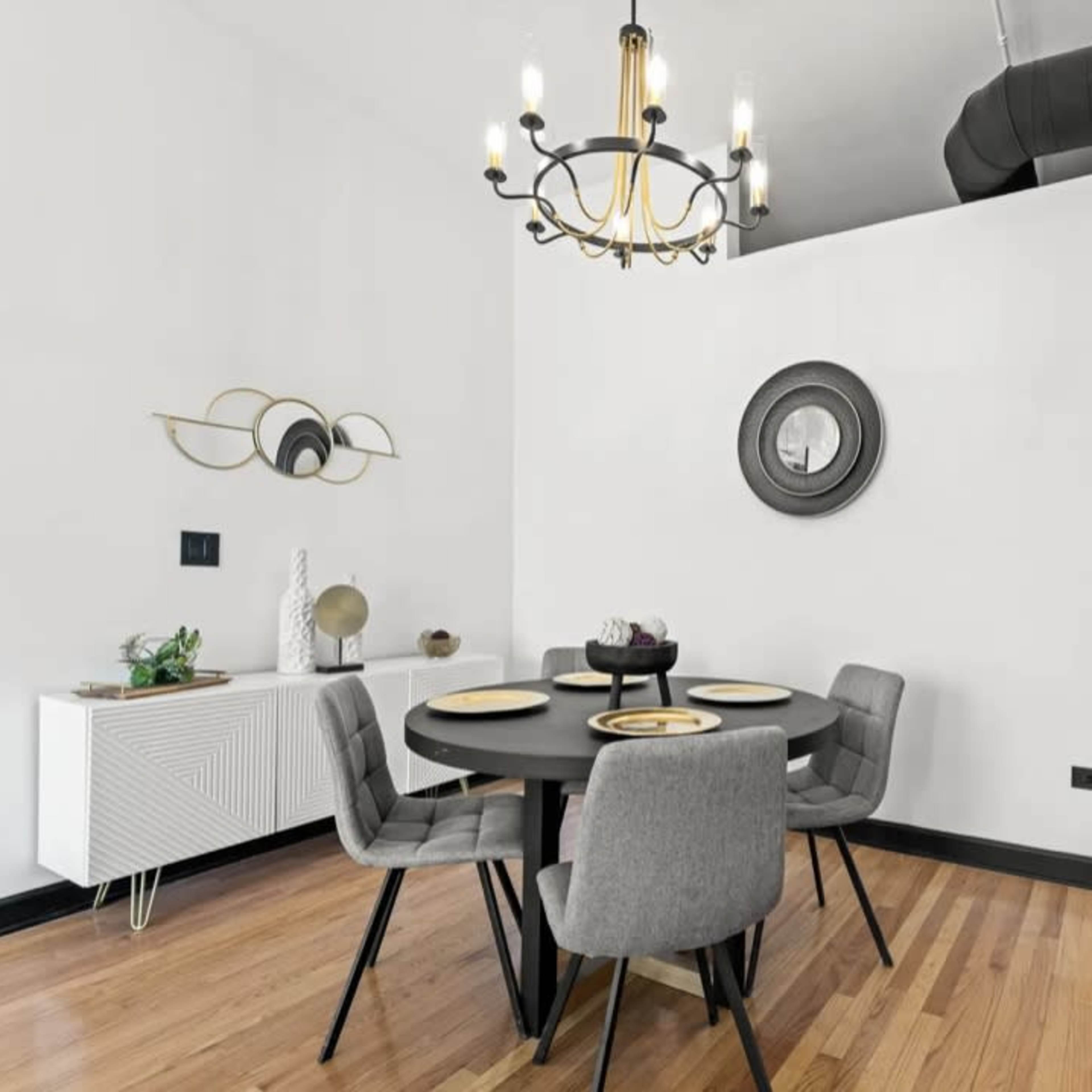 A modern dining area features a round black table set for four, surrounded by gray upholstered chairs, with a chandelier above and a sideboard against the wall.