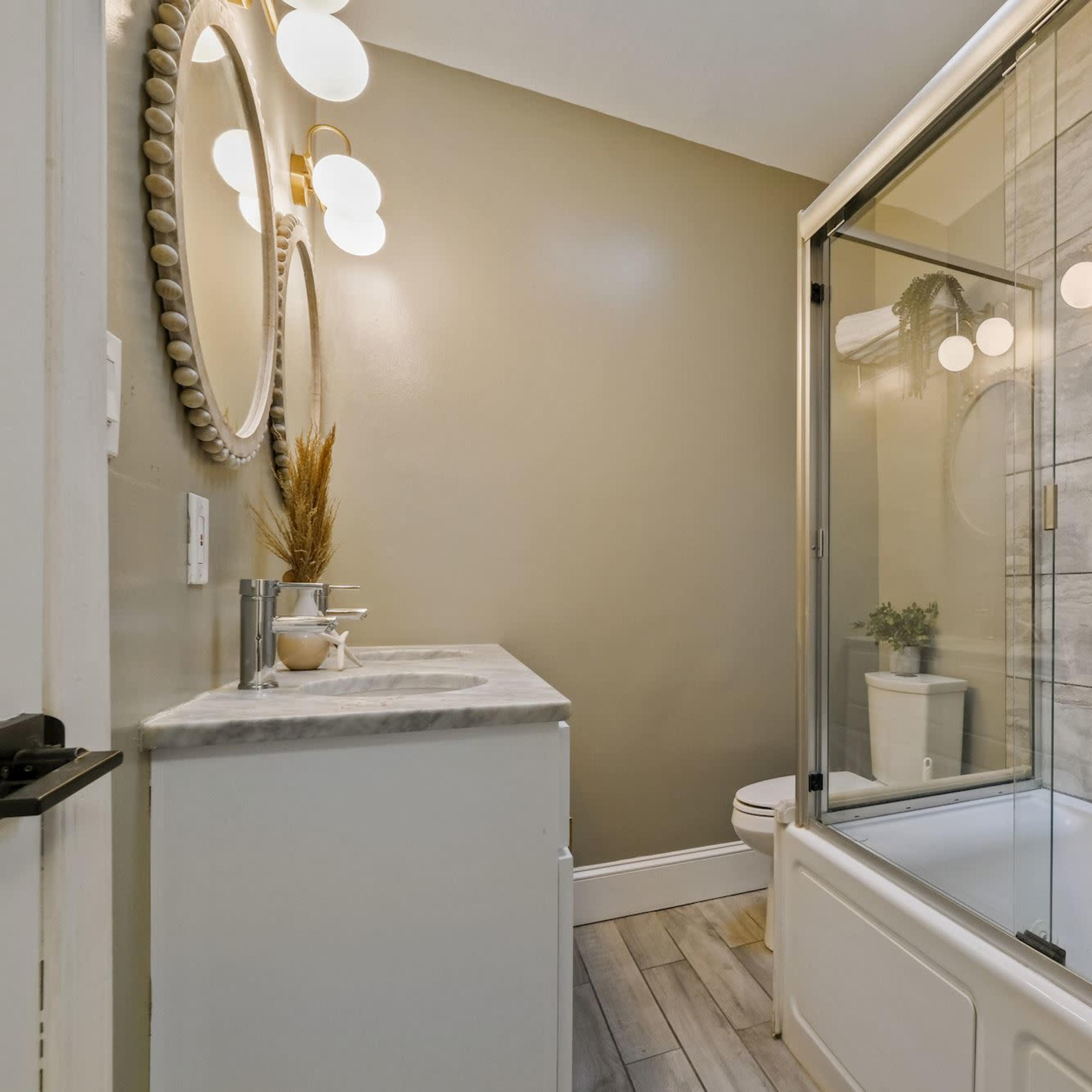 A well-lit bathroom featuring a glass shower enclosure, a white bathtub, and a vanity with a round mirror.