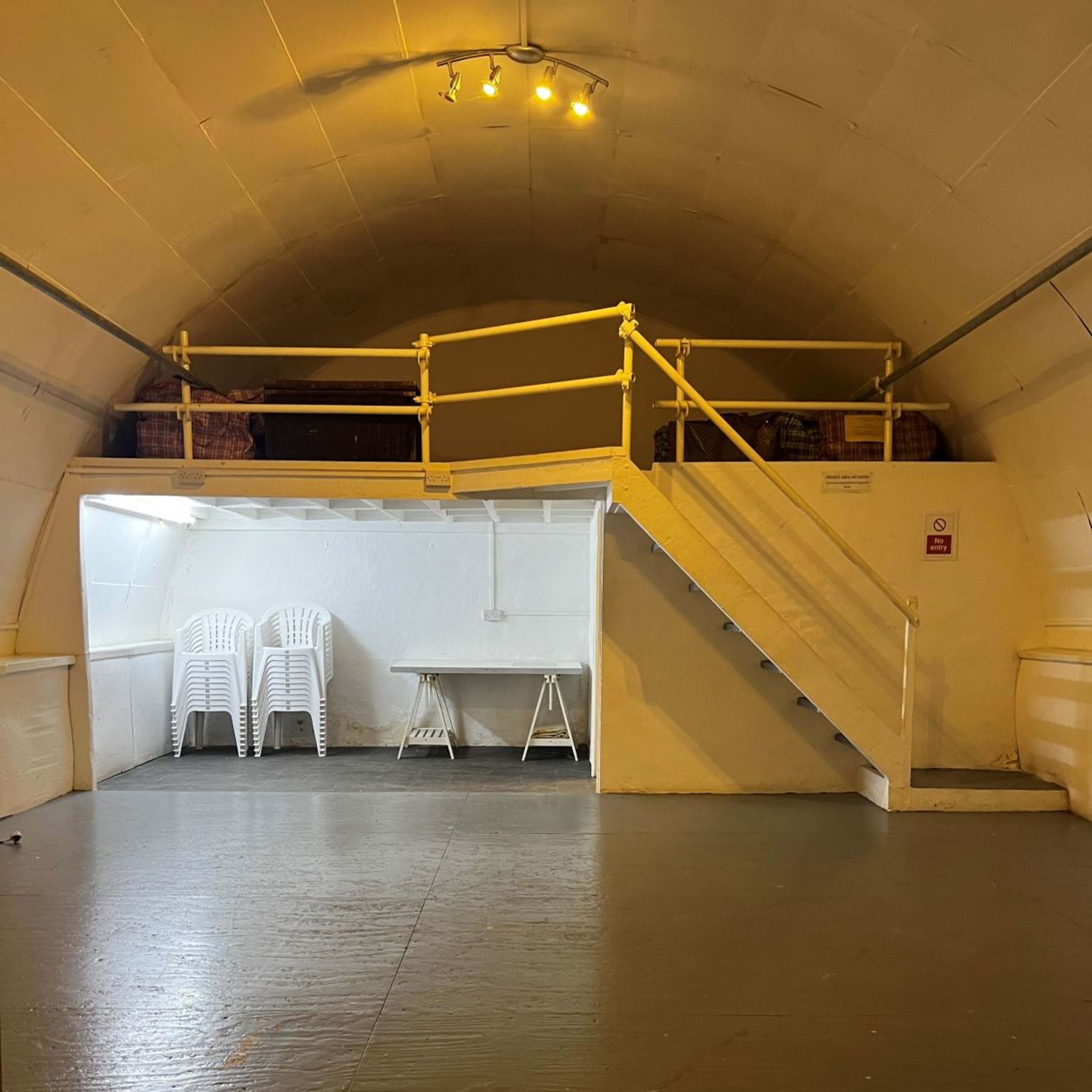 A spacious, well-lit bunker interior featuring a staircase leading to a raised platform with seating, and a table set up on the lower level.