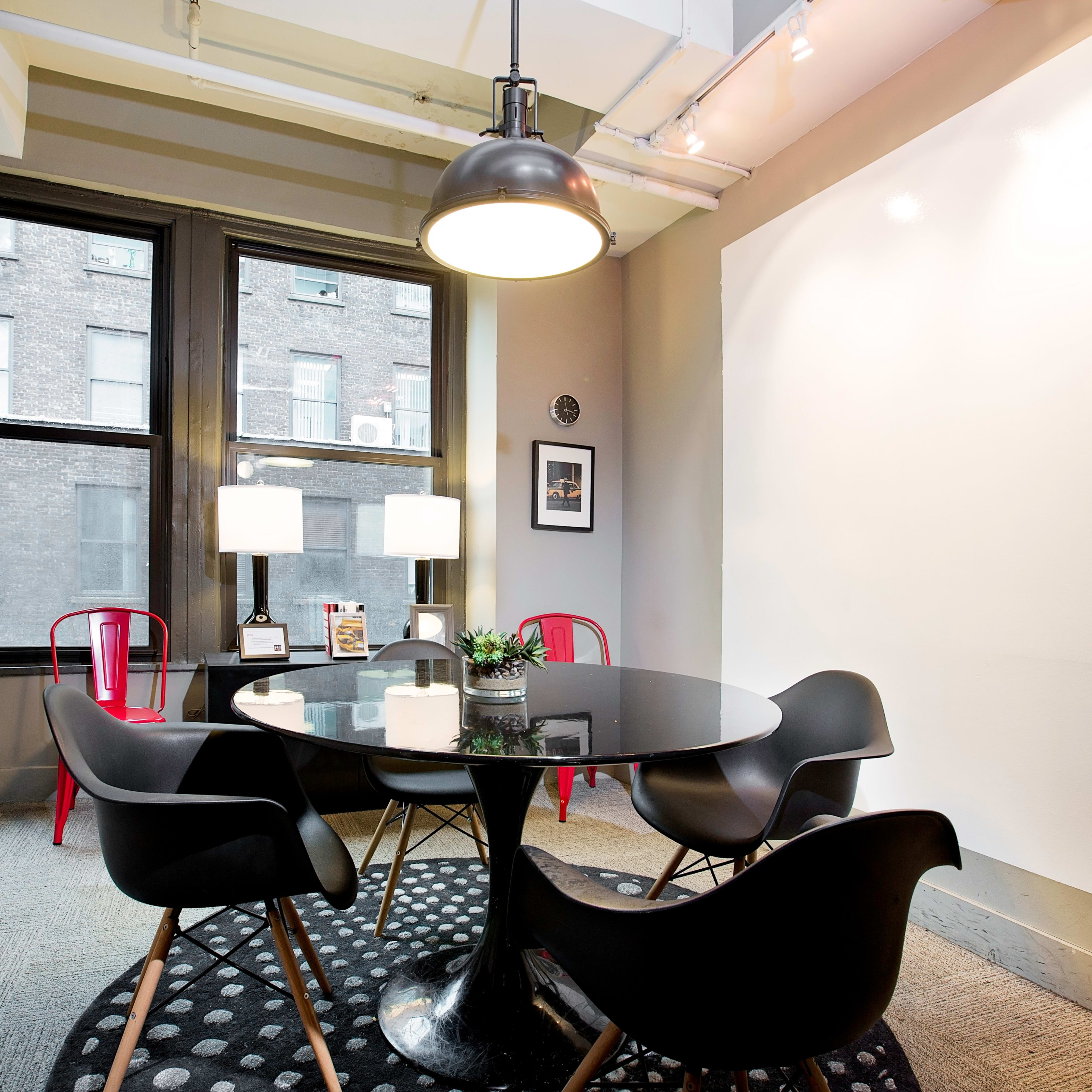 A modern small meeting room features a round black table surrounded by four chairs, with two lamps on a side table and framed pictures on the walls.