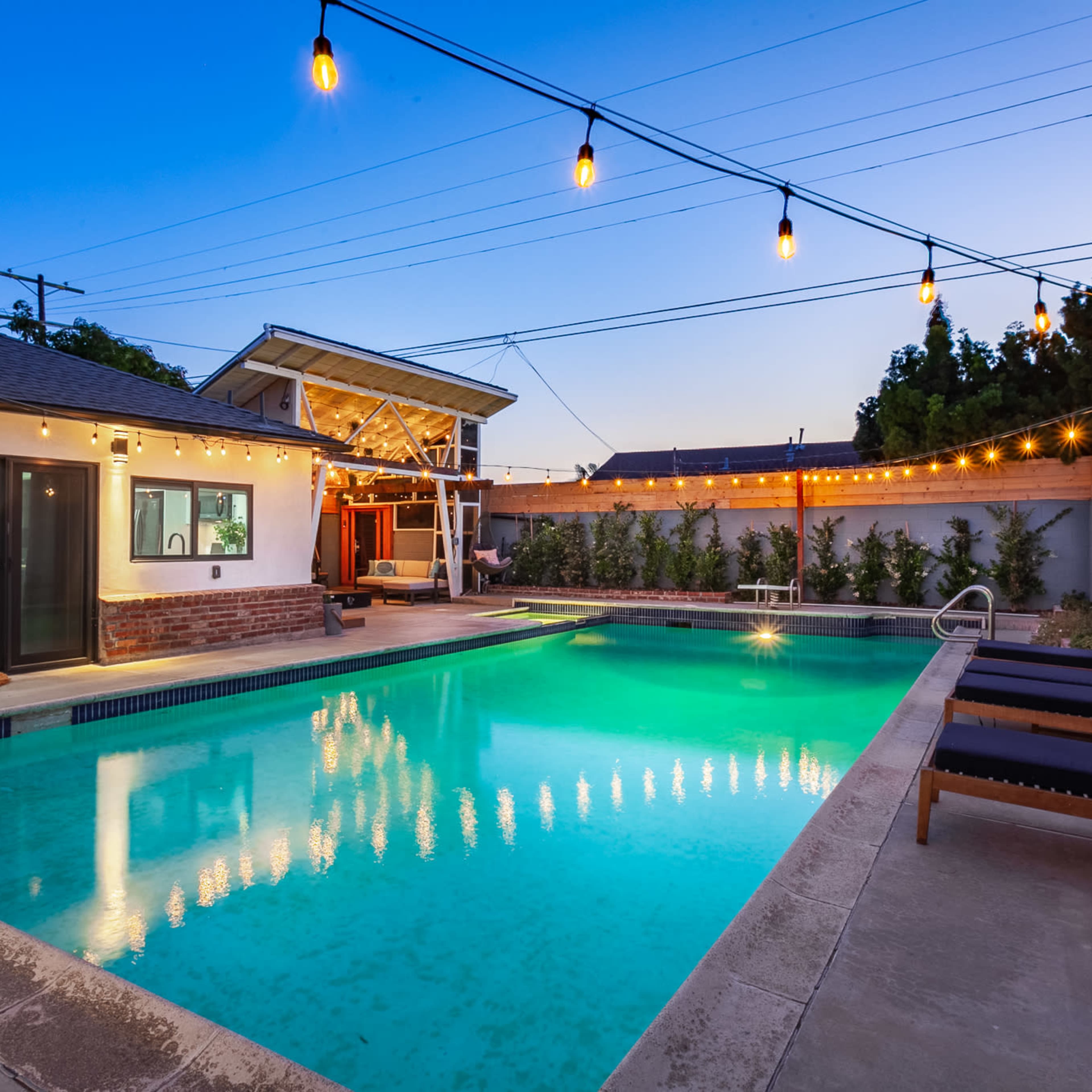 A well-lit backyard features a swimming pool surrounded by lounge chairs and string lights against a twilight sky.