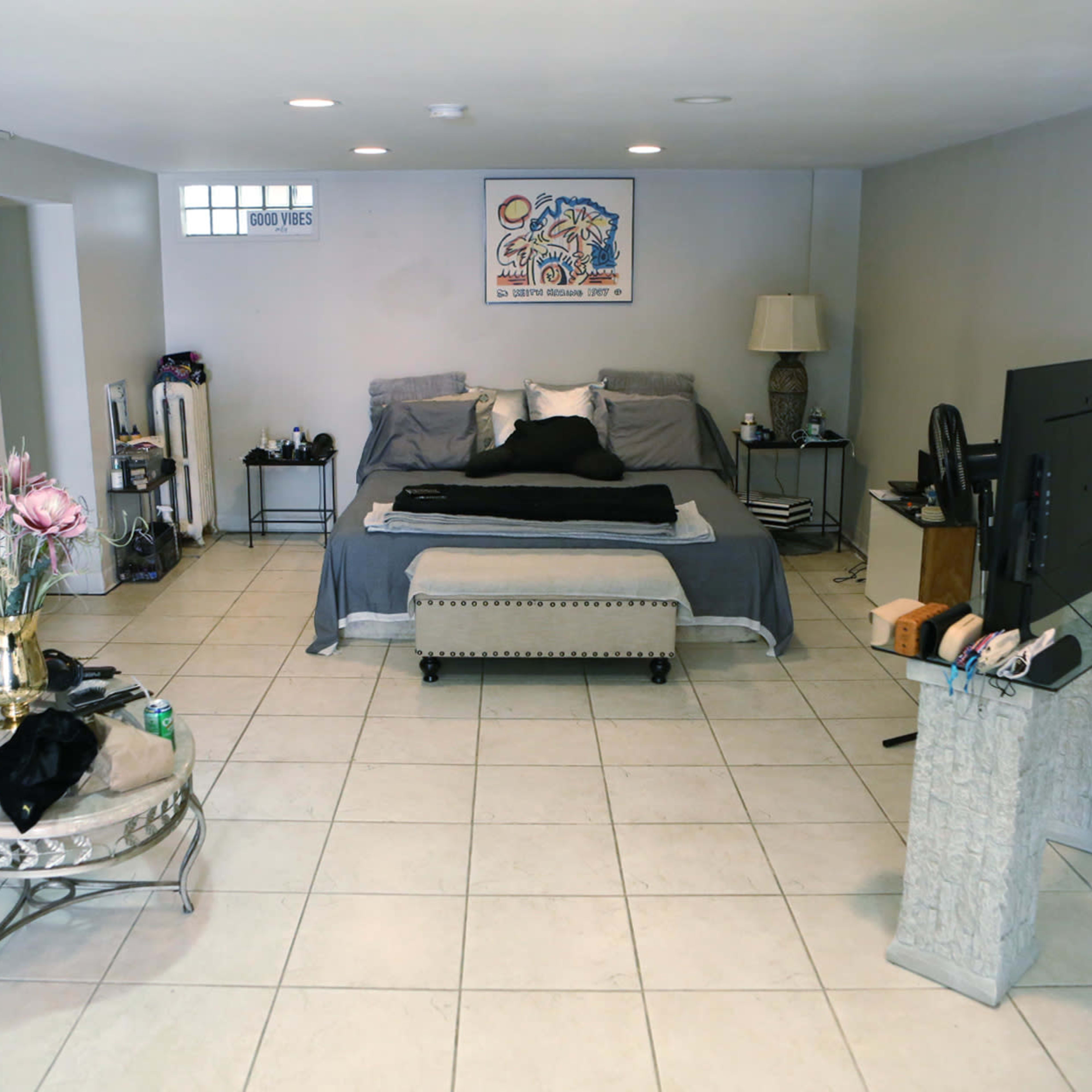 A spacious, minimalist bedroom with a bed at the center, a coffee table, and a television on a stand against a light-colored wall.