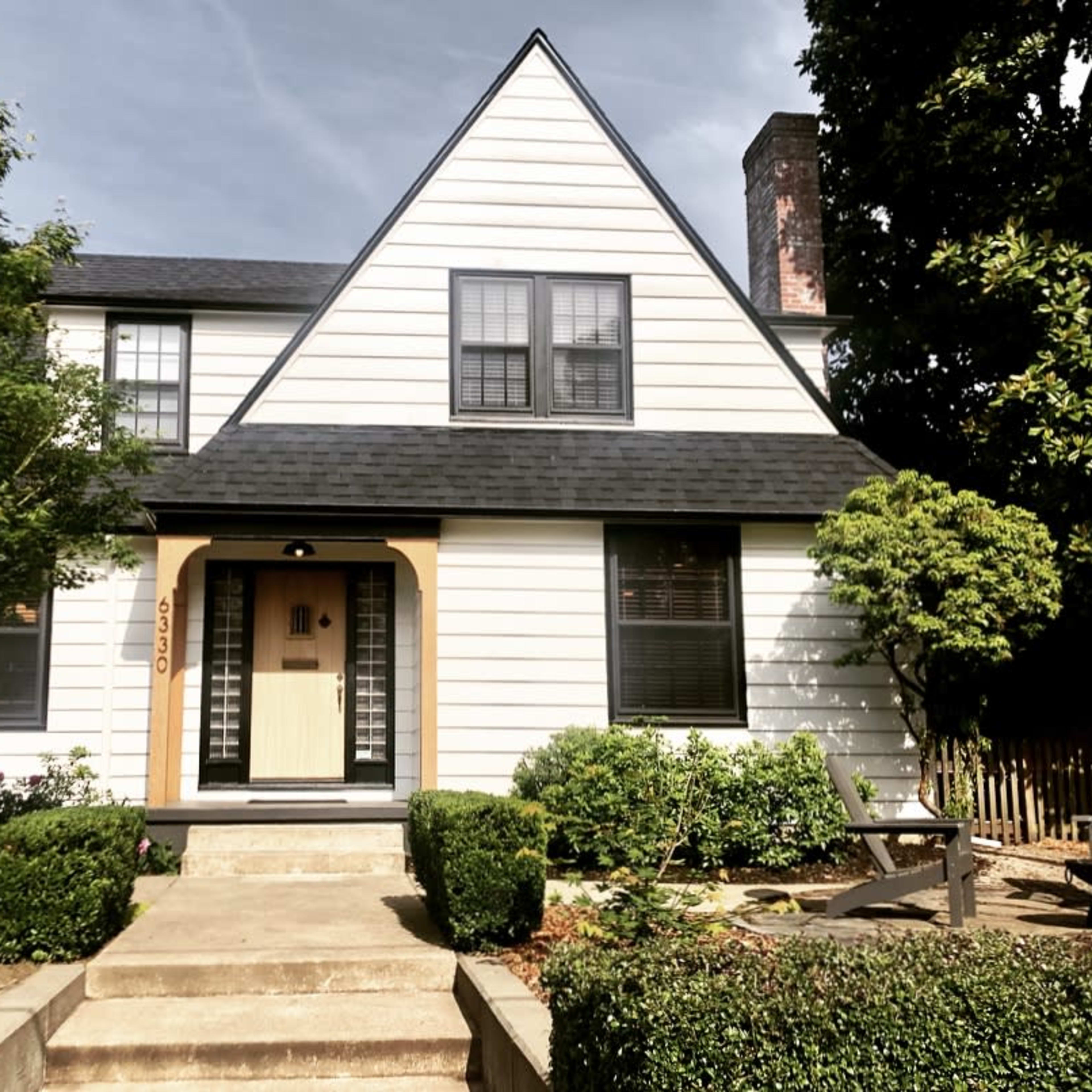 A two-story, A-frame style house with a white exterior, black roof, and a landscaped front yard.