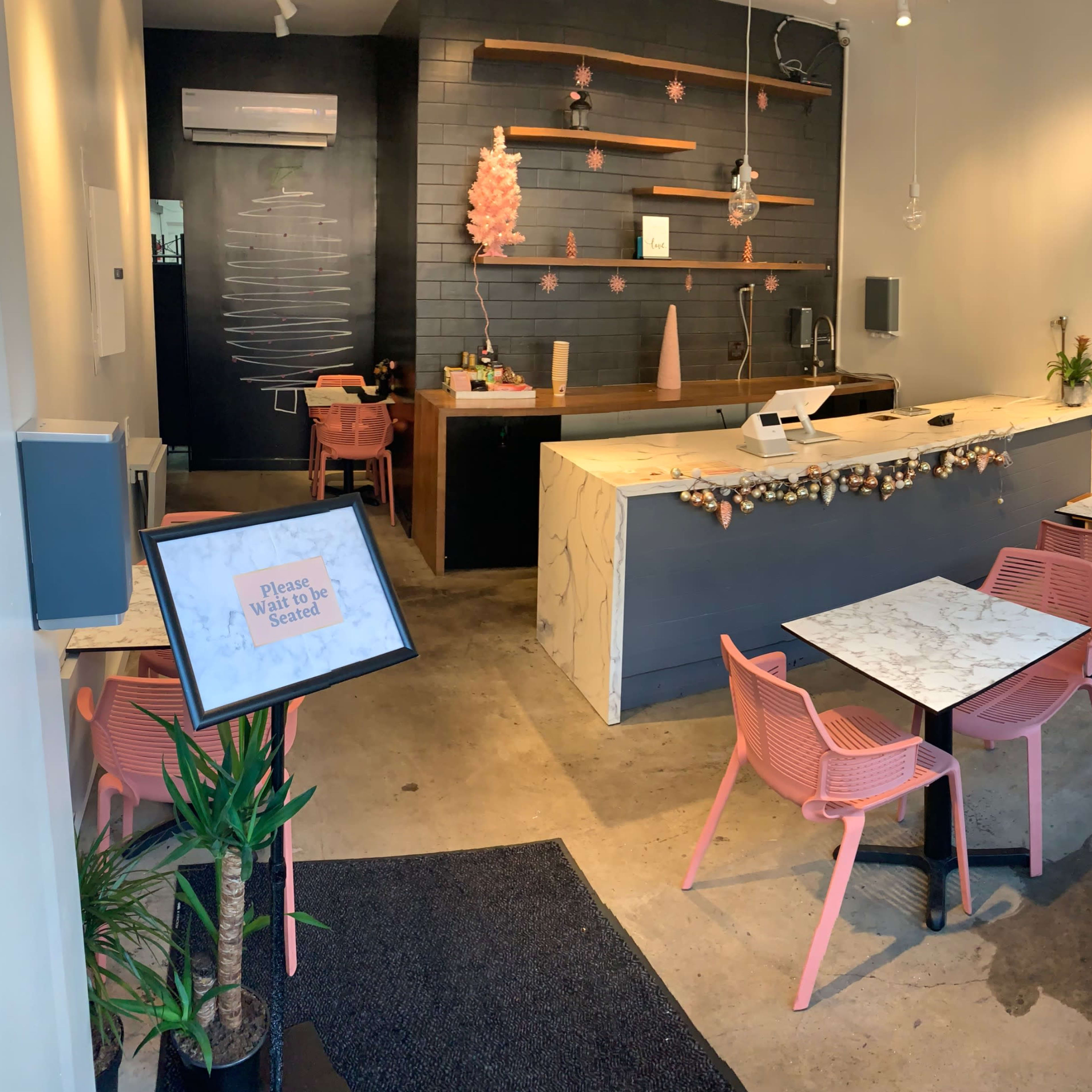 A modern cafe interior featuring marble-topped tables with pink chairs, a counter with decor, and plants in the corners.