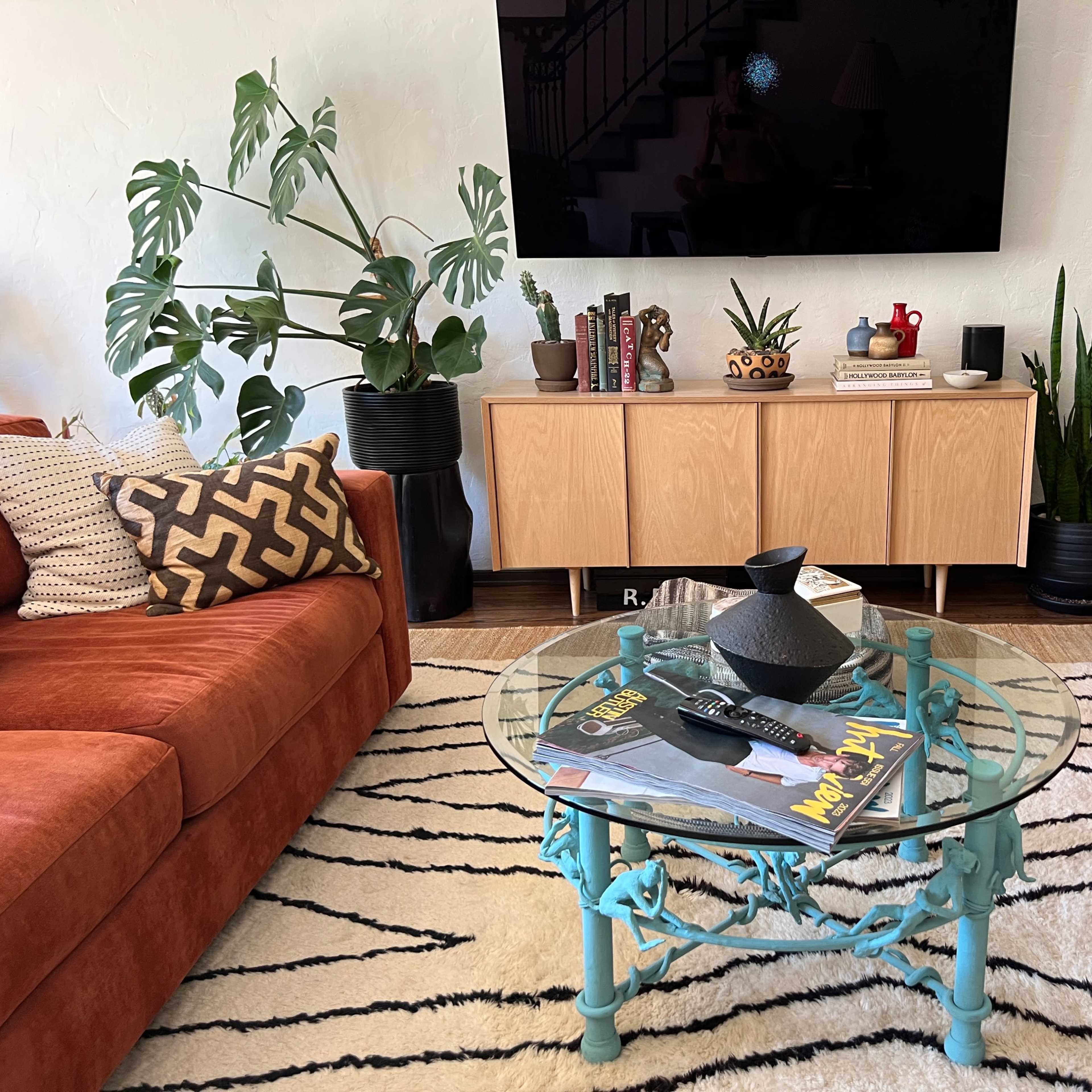 A living room features two orange sofas flanking a circular glass coffee table on a striped rug, with a television mounted on a light-colored wall.