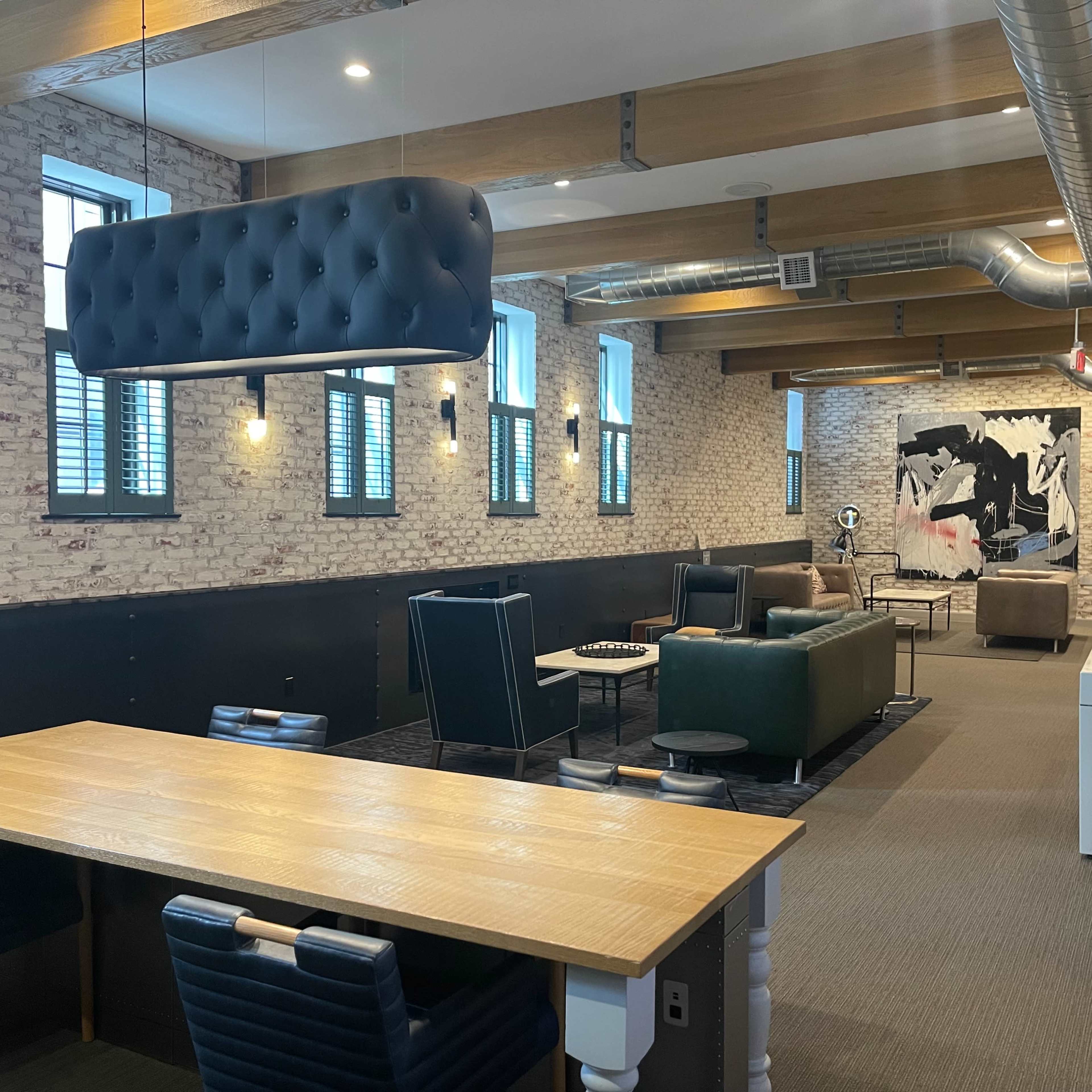 A spacious lounge area with exposed brick walls, seating arrangements including sofas and chairs, and a large wooden table.