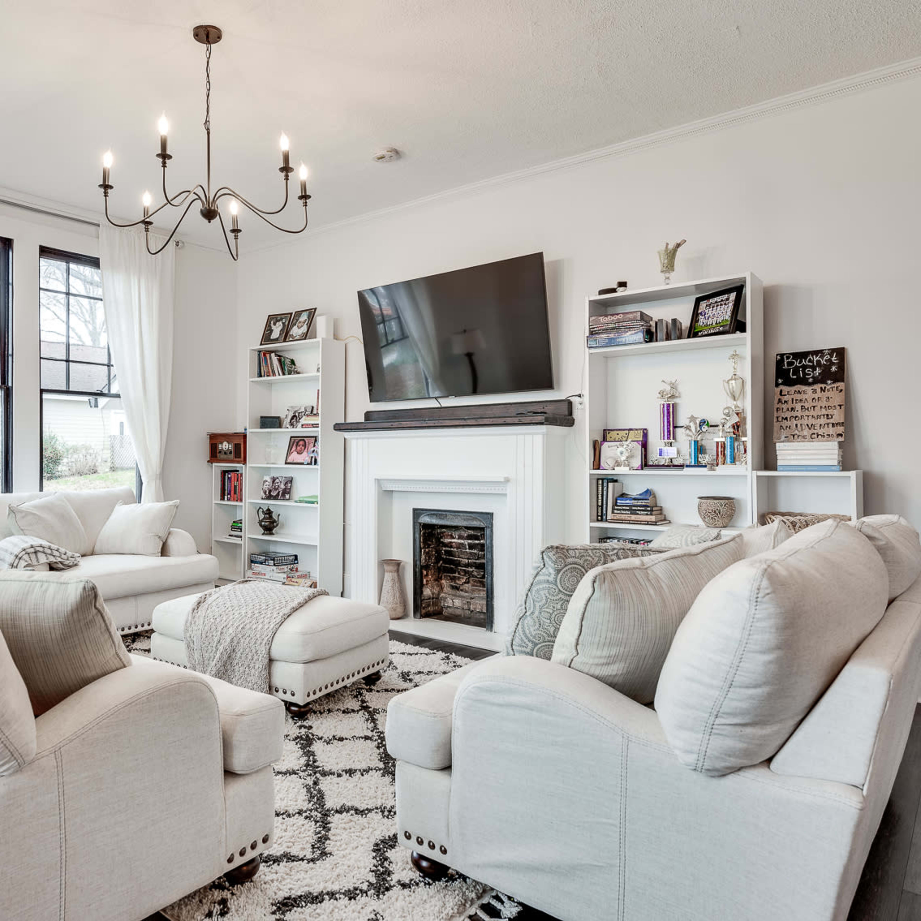 The living room features a chandelier, a large television mounted above a fireplace, and white sofas arranged around a patterned rug.