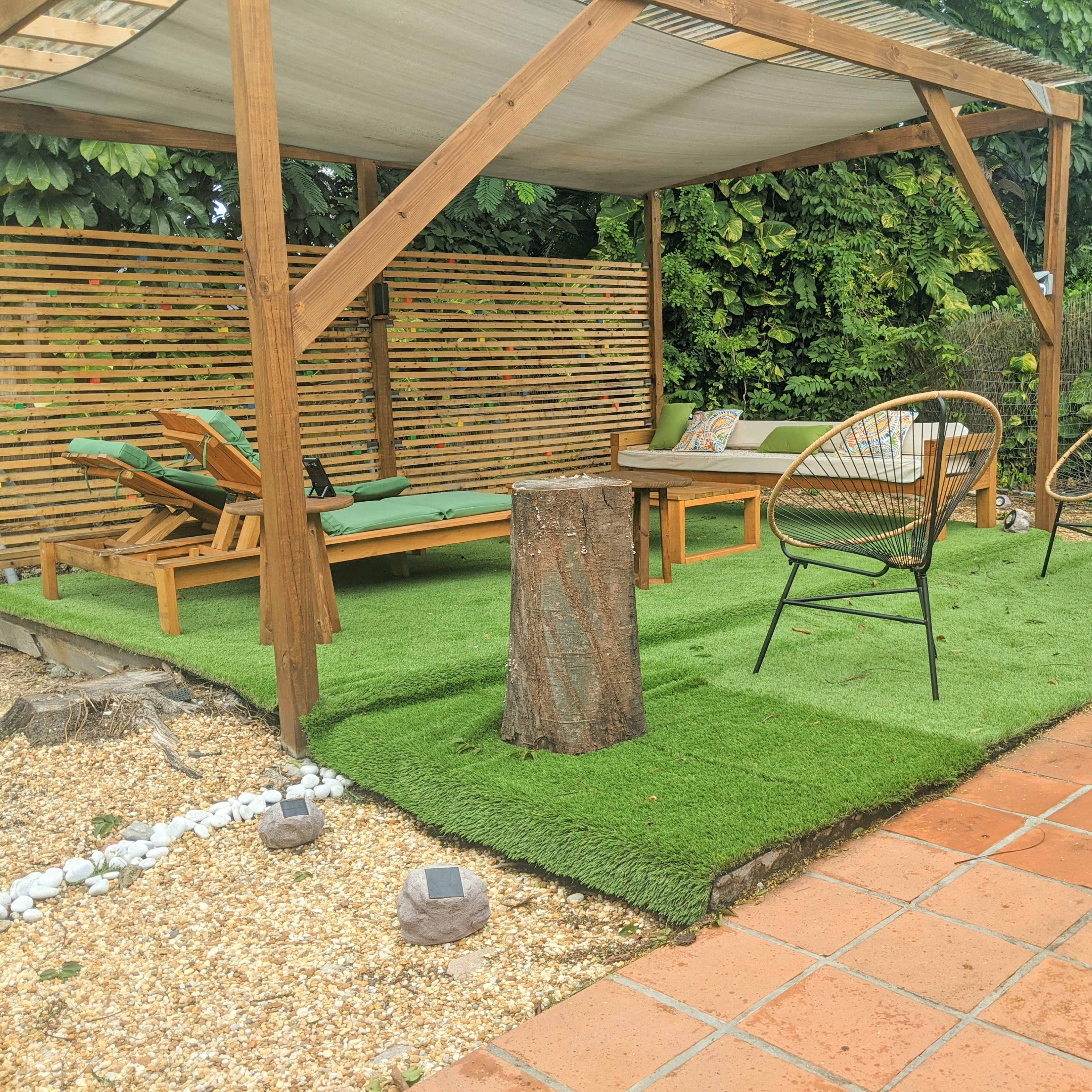 A shaded outdoor seating area features a wooden pergola, two metal chairs, a lounge chair, and a tree stump on artificial grass with gravel and stone accents.