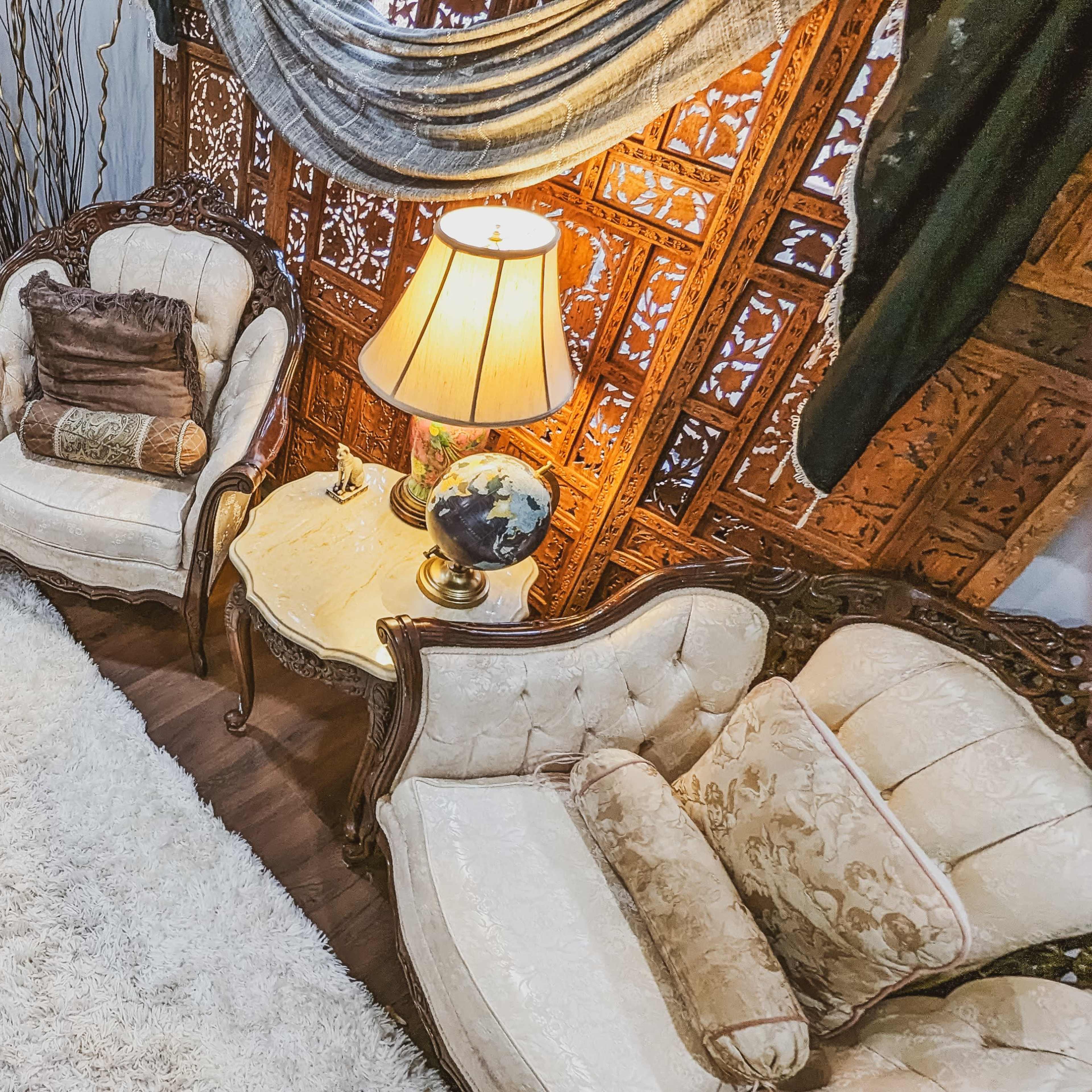 A cozy living room with ornate upholstered furniture arranged around a central coffee table on a plush rug, illuminated by a lamp next to an intricately carved wooden screen.