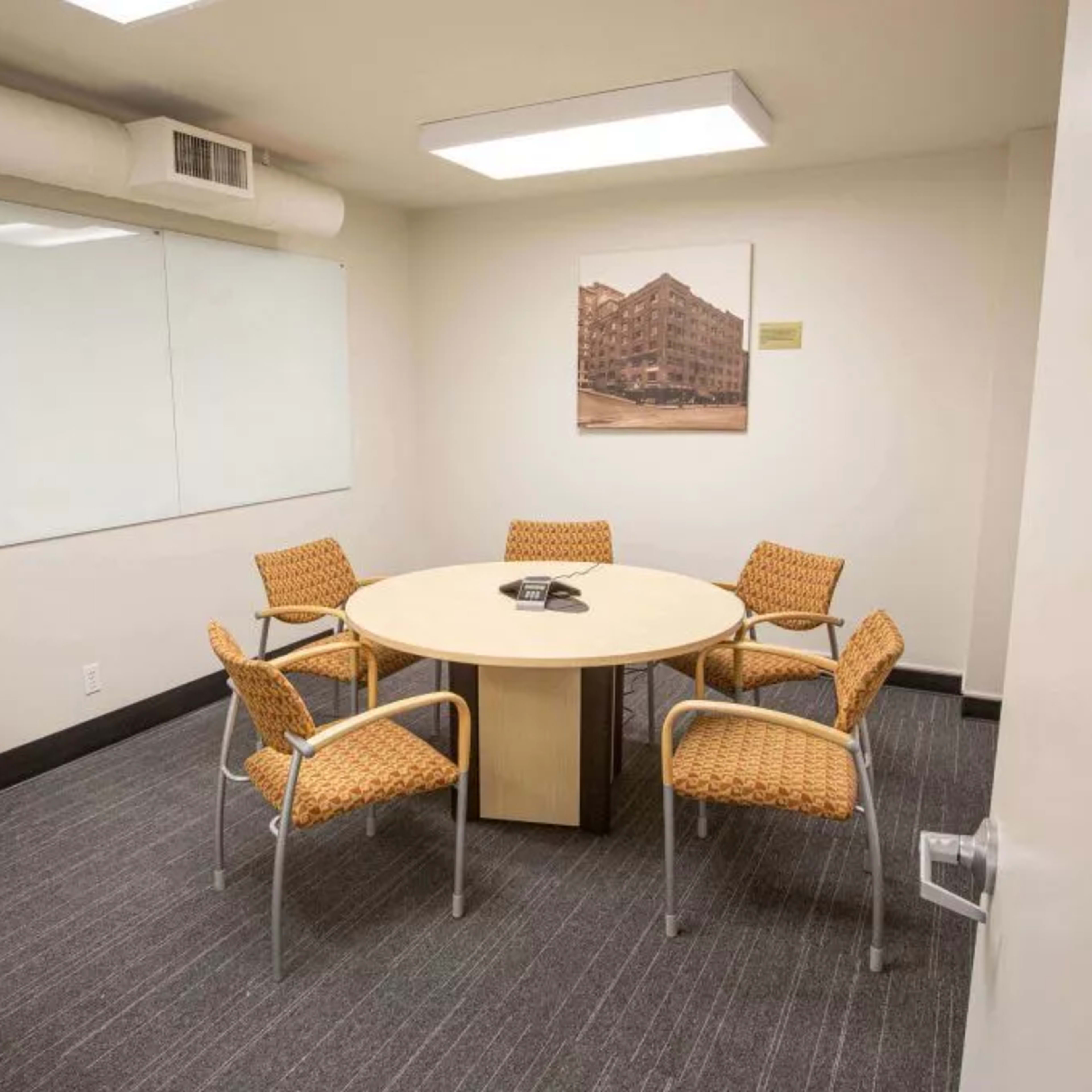 A small meeting room with a circular table surrounded by four orange chairs, a whiteboard on the wall, and a wastebasket nearby.